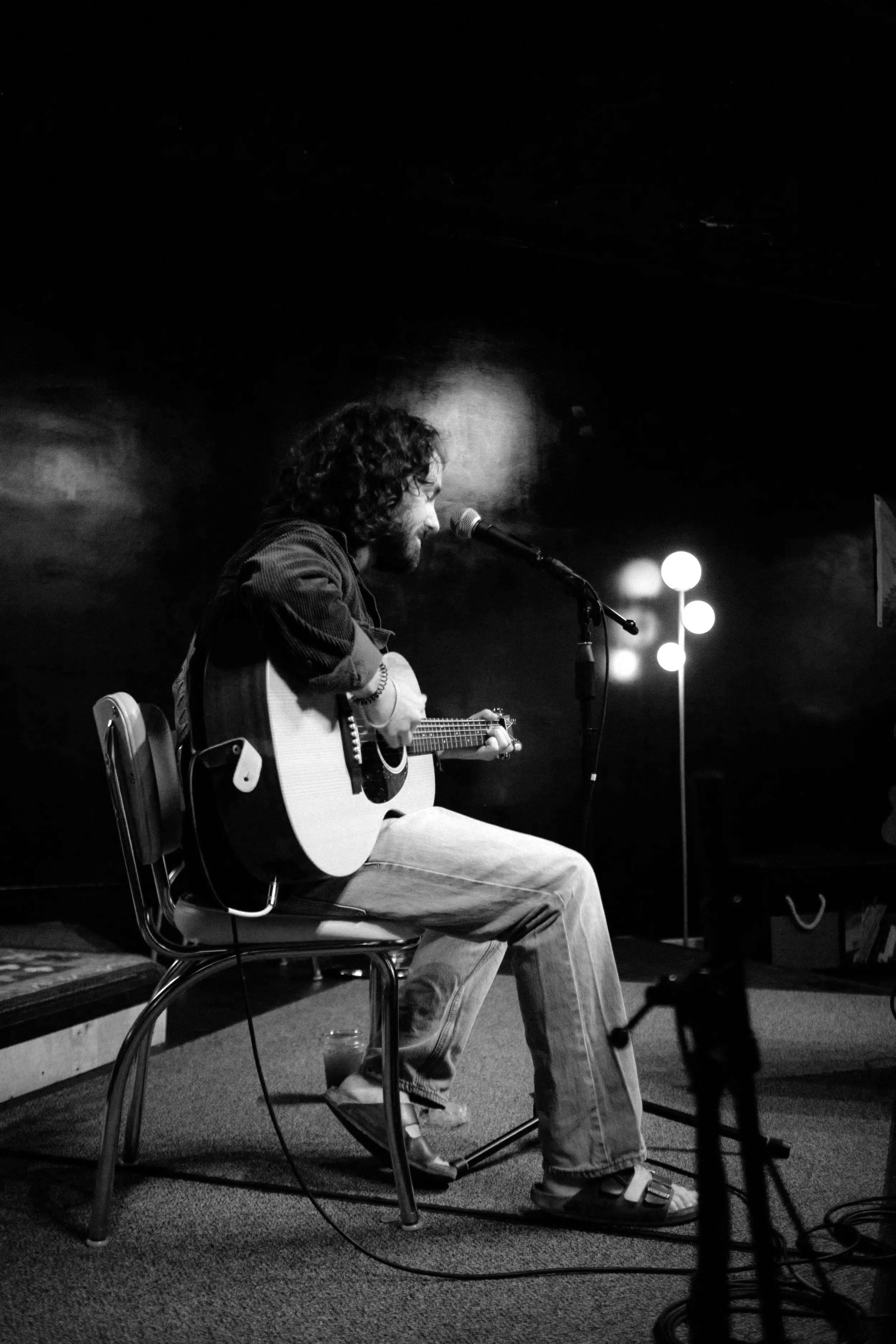 Witness Trips, a musician with long curly hair and glasses sitting on a chair and playing an acoustic guitar, singing into a microphone in a dimly lit room.