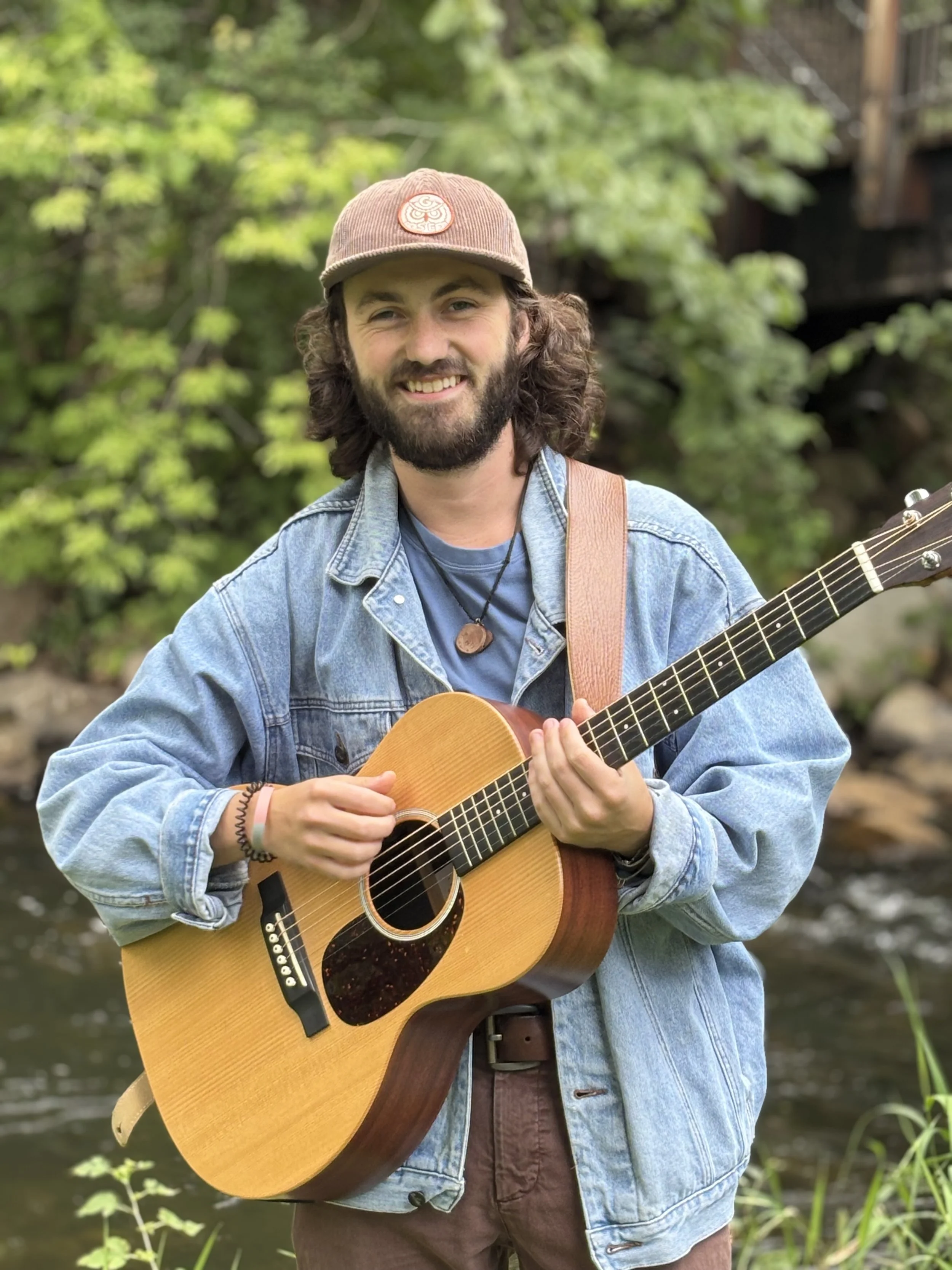 A young man with long dark hair and a beard standing outdoors, holding an acoustic guitar, smiling, wearing a brown cap, denim jacket, and brown pants, with trees and a river in the background.