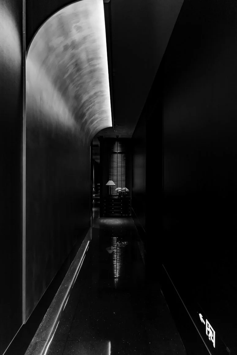 A dimly lit, narrow corridor with dark walls and a shiny black floor, reflecting light. At the end of the corridor, there are window blinds and a hanging light fixture over a small table with decorative items.