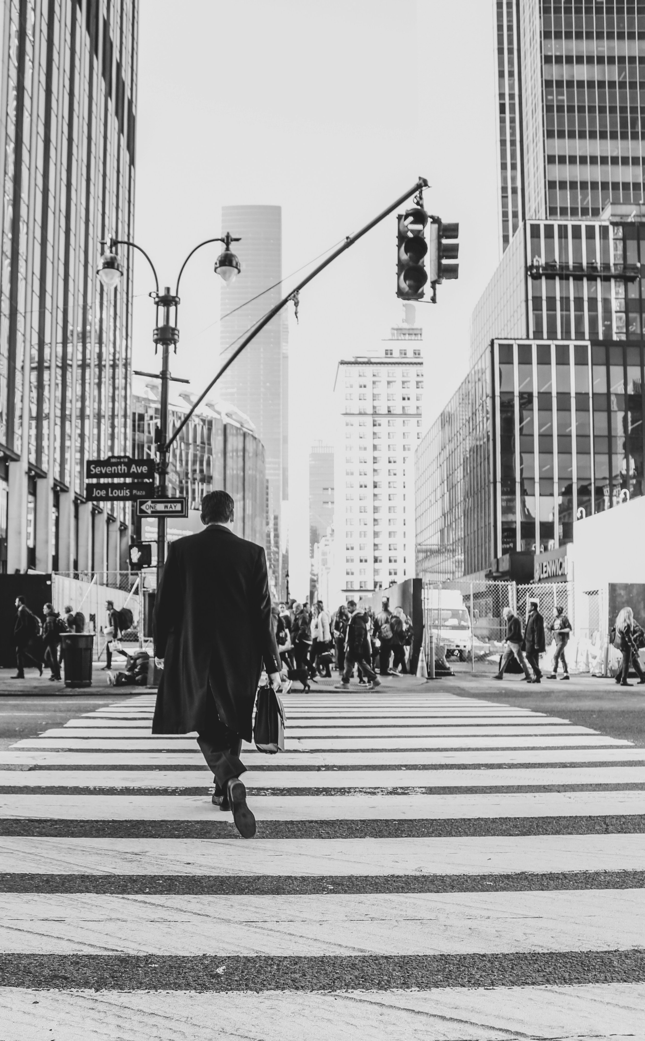 Black and white photo of a man in a coat carrying a bag walking across a city crosswalk with tall buildings and a traffic light overhead, in an urban downtown area.