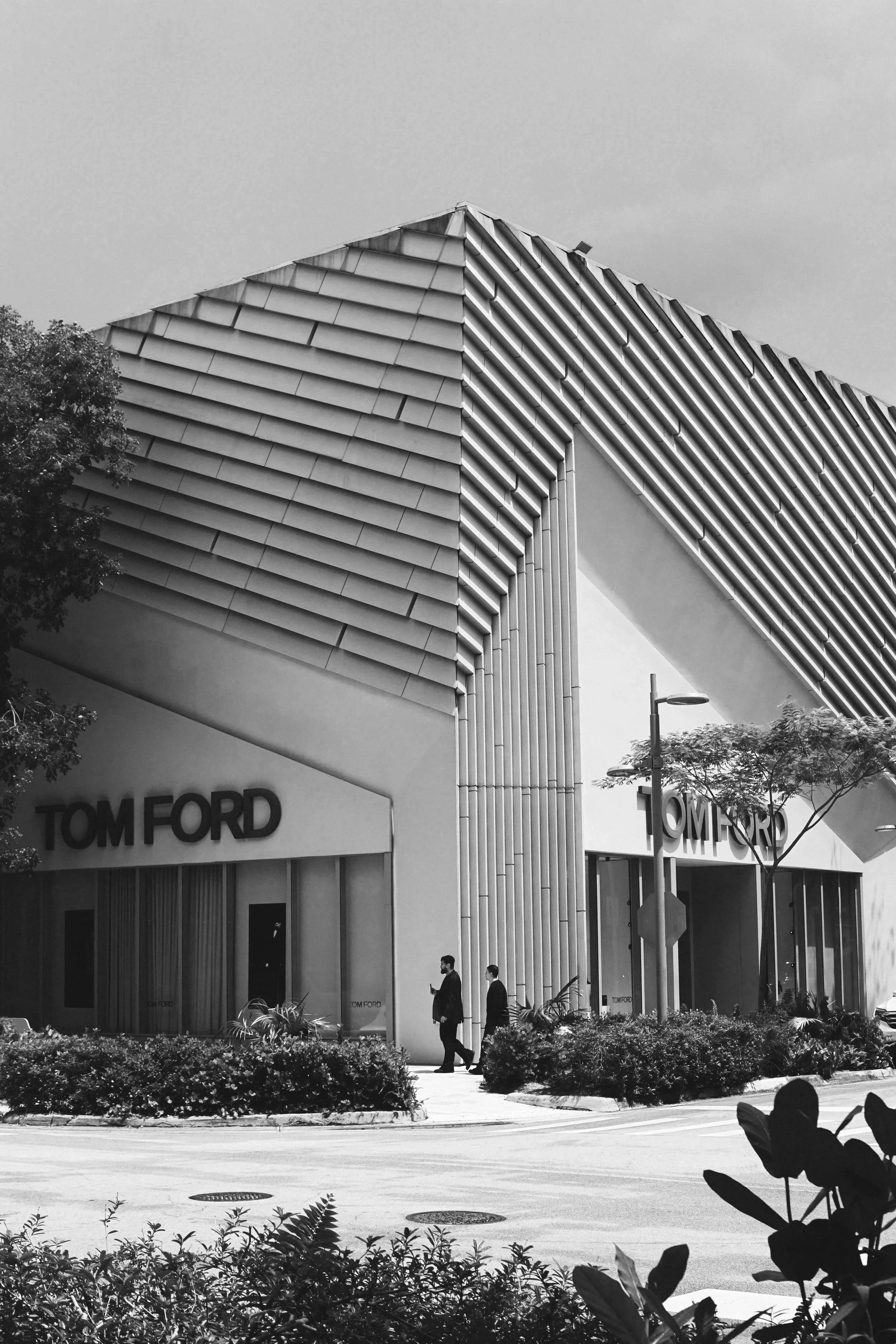 Exterior view of a modern Tom Ford store with large signage, featuring a contemporary architectural design with horizontal lines and geometric shapes, with pedestrians walking by.