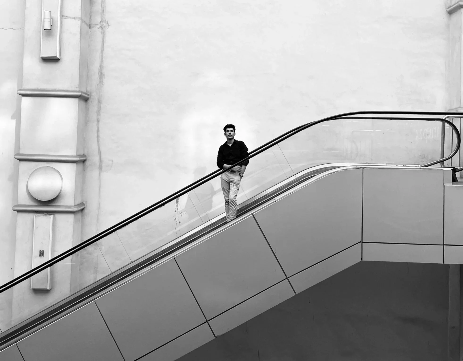 A young man standing on an indoor escalator in front of a plain wall, dressed in a dark shirt and light pants, in black and white photo.