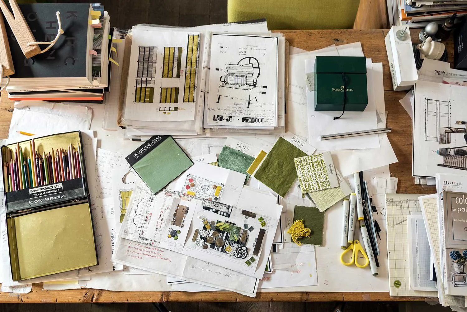 A cluttered wooden desk with architectural drawings, color swatches, colored pencils, pens, scissors, and notes related to interior design or architecture.