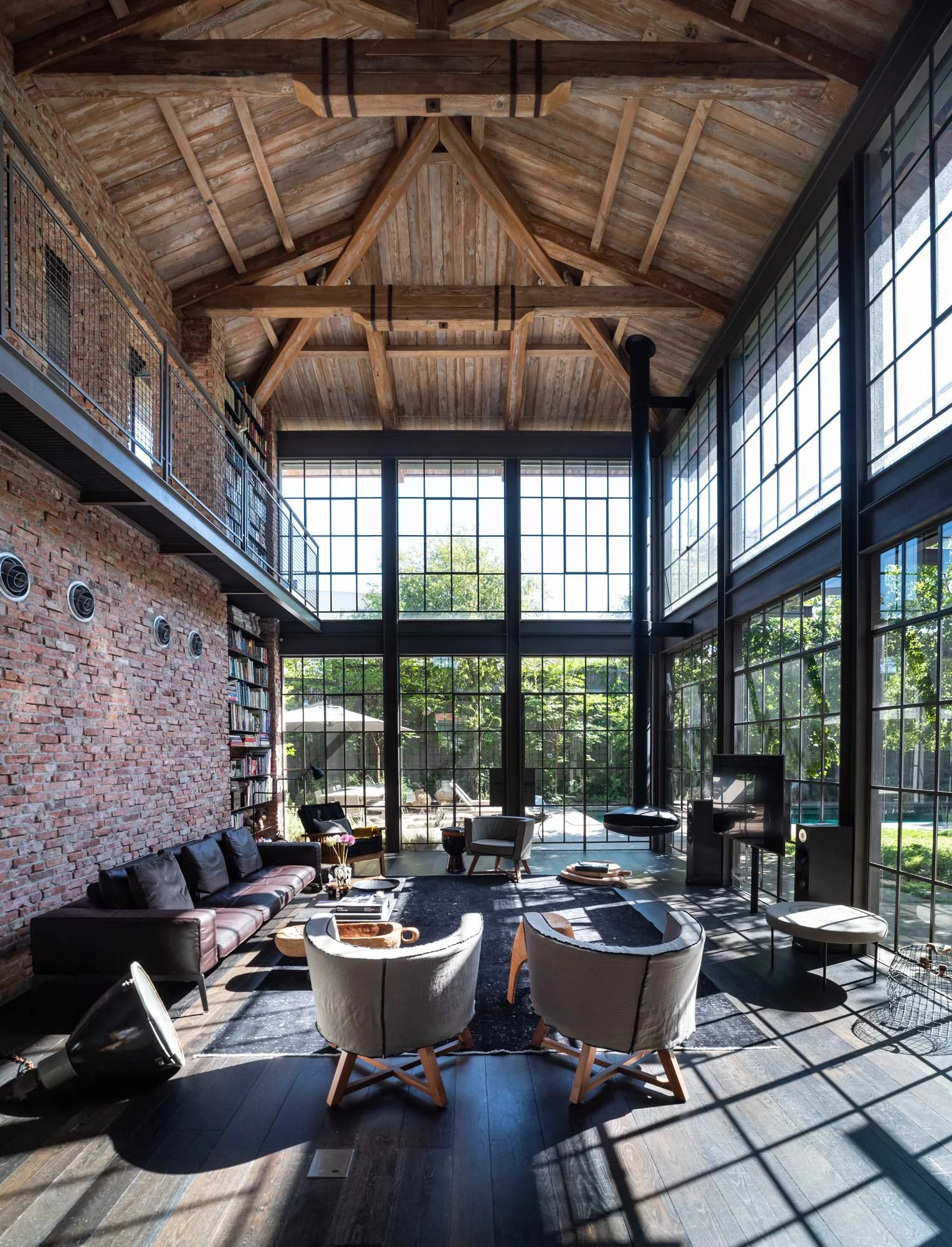 Interior view of a spacious living room with large windows, exposed brick walls, a high wooden ceiling with beams, and modern furniture including armchairs and a leather sofa, with sunlight casting shadows.