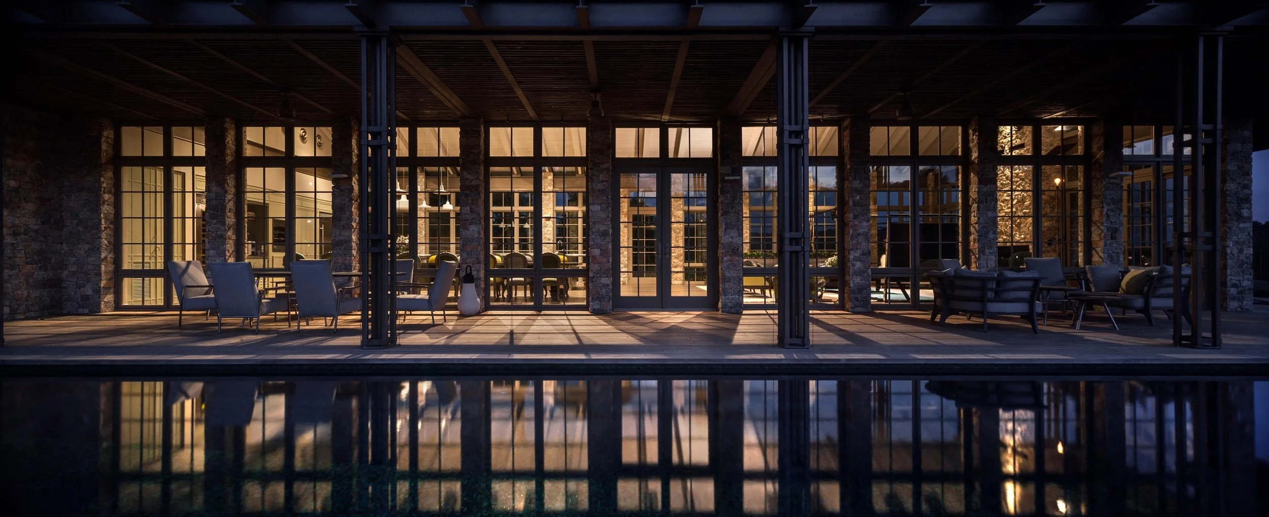 Exterior view of a modern house with large glass windows and a patio area, illuminated from inside, with a swimming pool in the foreground at night.
