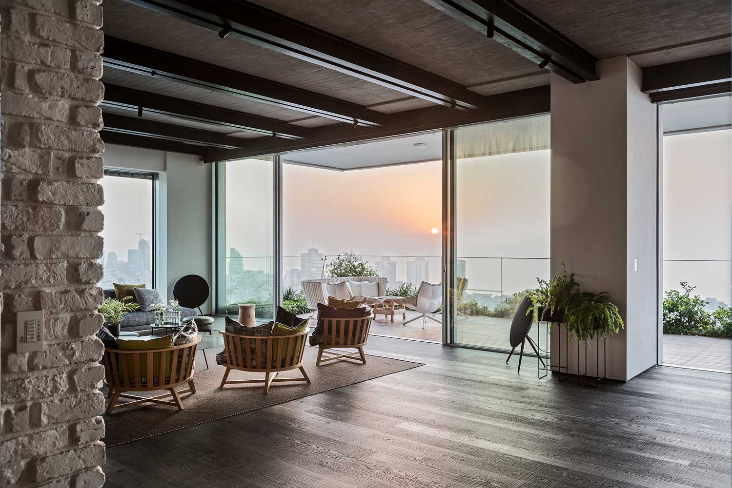 Modern living room with large glass windows and balcony, featuring outdoor seating and city skyline view at sunset.