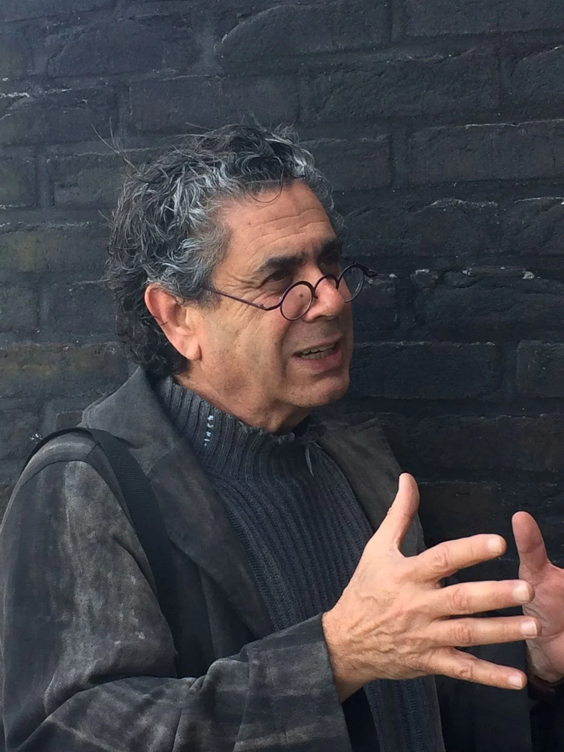 A man with gray curly hair, wearing glasses, a gray jacket and a dark sweater, is standing against a dark brick wall. He appears to be explaining something, using hand gestures.