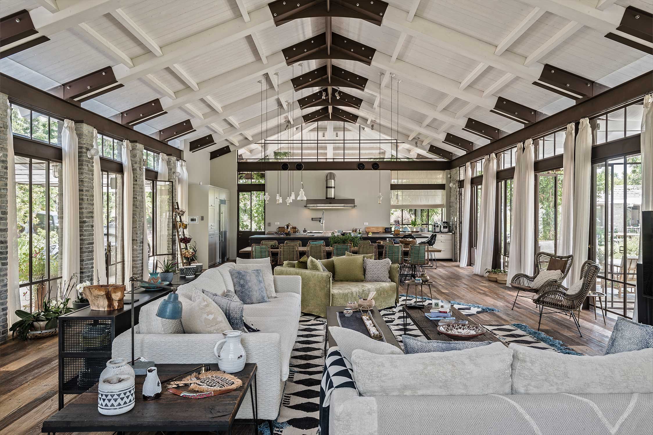 An open-concept living room and kitchen with high vaulted ceiling, exposed rafters, and large windows with white curtains, decorated in a modern rustic style.