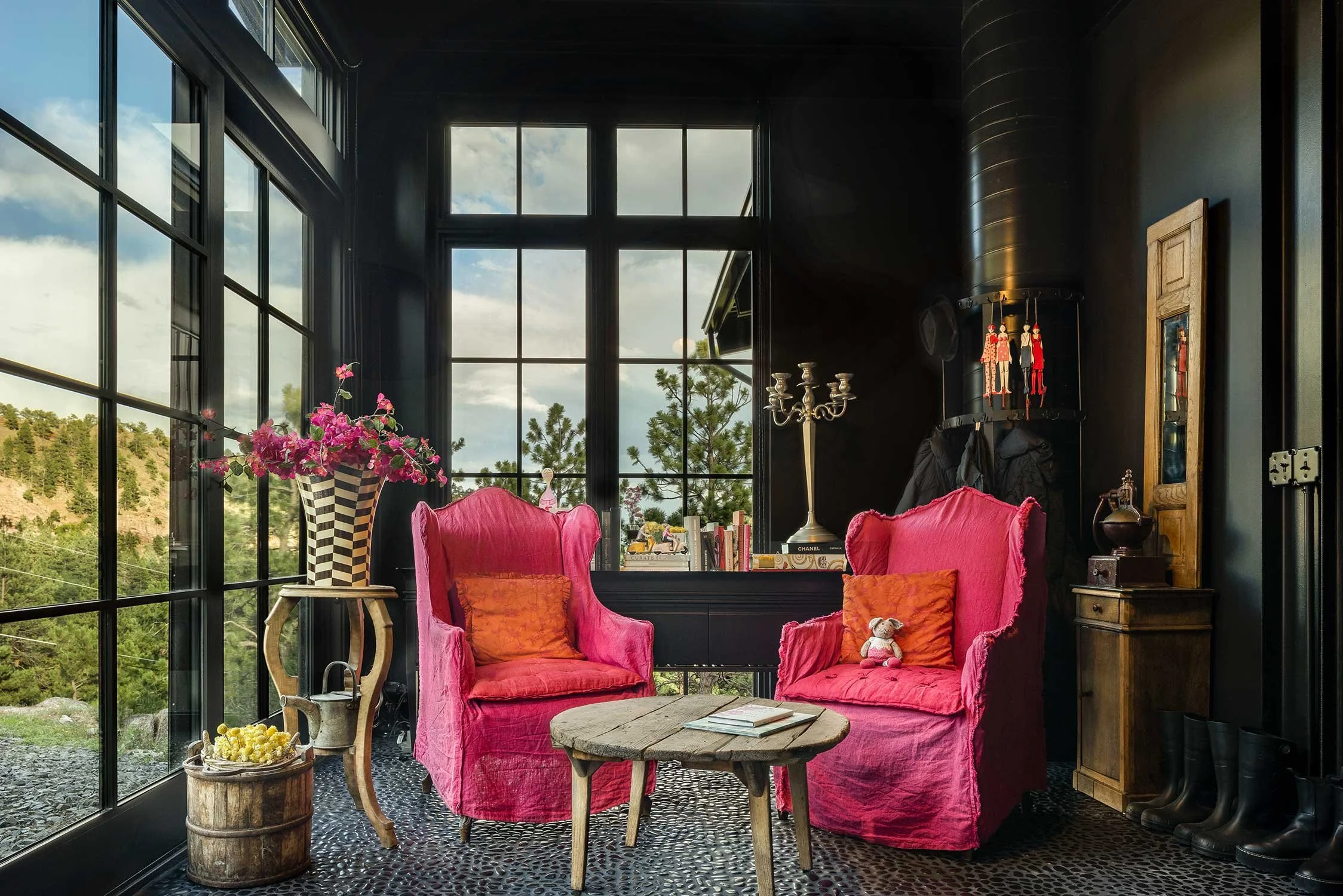Interior of a room with large black-framed windows showing a view of green hills and a cloudy sky, decorated with pink velvet armchairs, a small rustic wooden table, a tall candelabrum, a wooden cabinet, and various decorative items including a vase with pink flowers.
