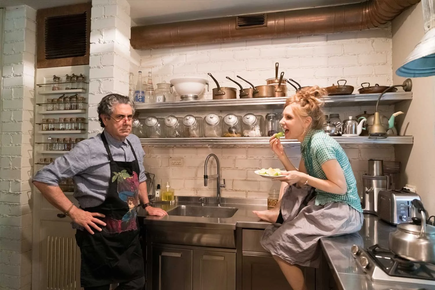 A man with gray hair and glasses, wearing a gray shirt and apron, stands with hands on hips, facing a woman who is sitting on the counter eating a plate of food. The woman has curly red hair and is wearing a green checkered shirt and a gray skirt. They are in a kitchen with white brick walls, shelves with jars and cookware, a sink, and various kitchen appliances.
