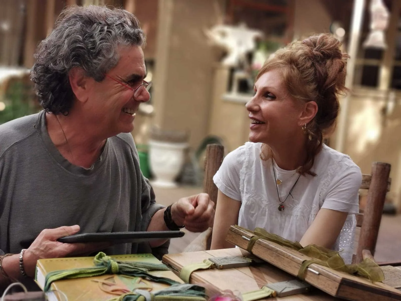 A man and a woman sitting at a table, smiling at each other and engaging in conversation, in a cozy indoor setting with warm lighting and blurred background.