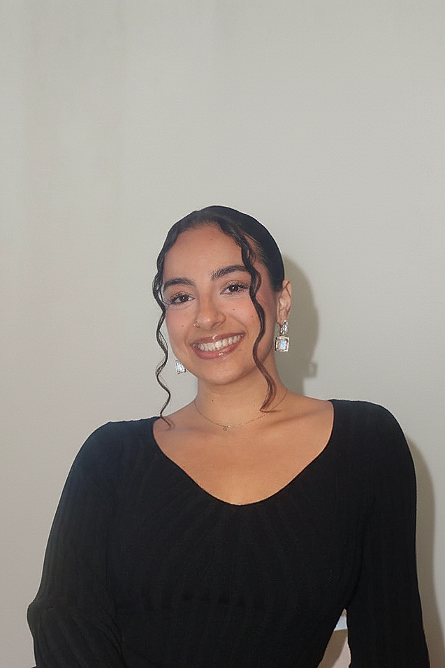 A young woman with dark, curly hair and light brown skin smiling, wearing a black dress and silver dangling earrings, standing against a plain white wall.