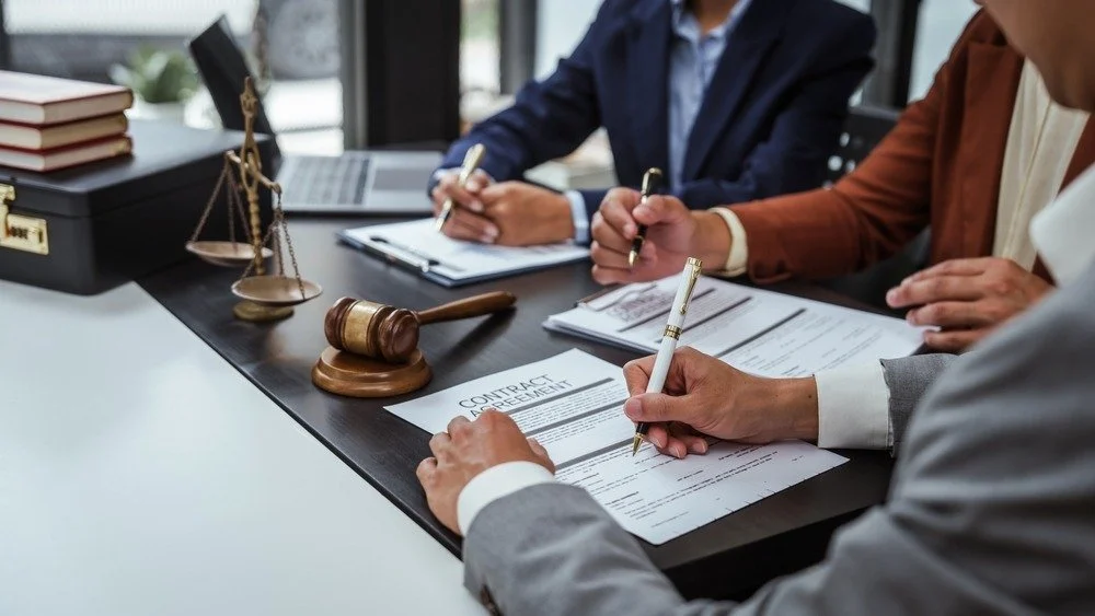 People in business attire signing a contract at a desk with legal books, a gavel, and a scale of justice regarding business law dispute.