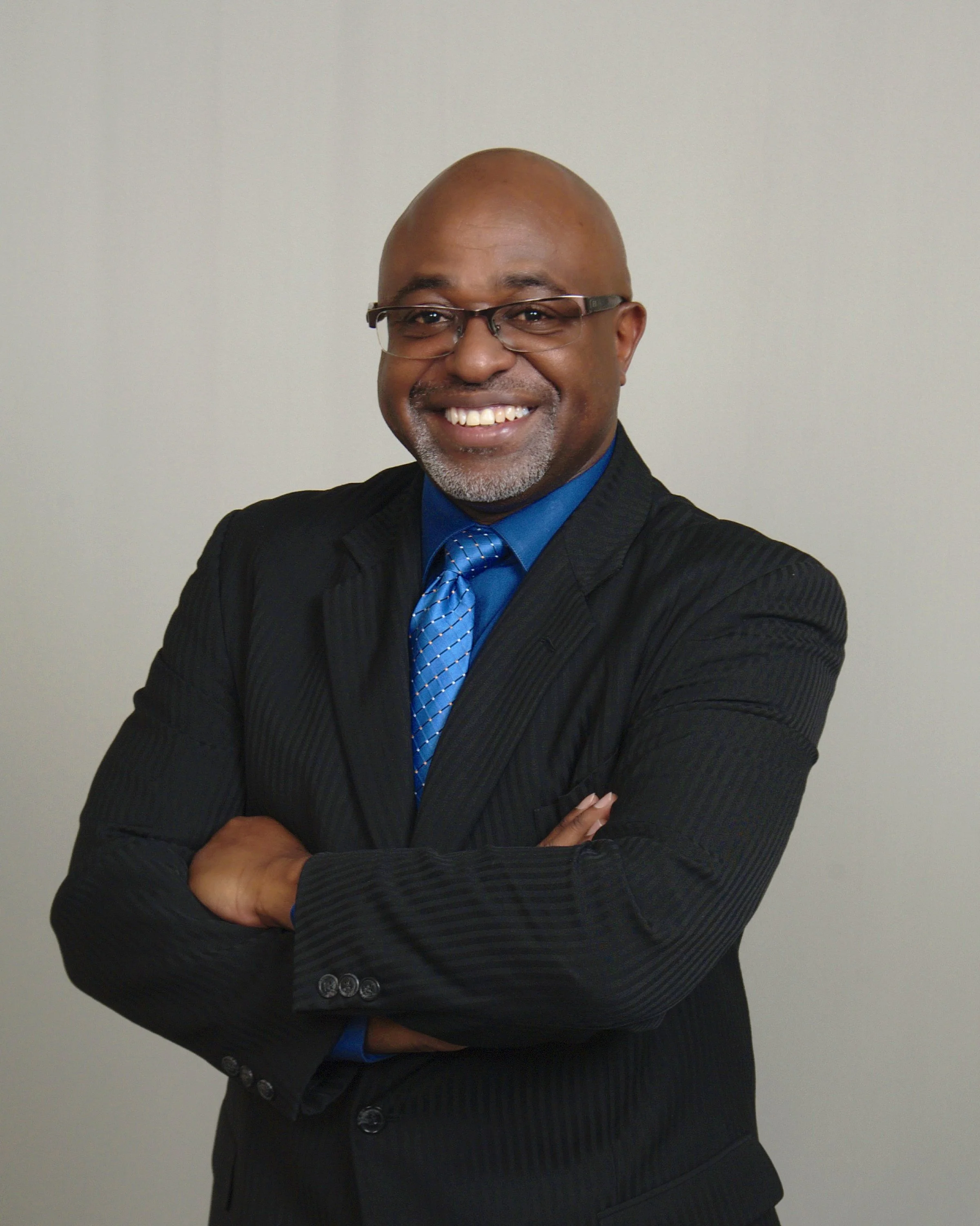 A smiling African American man wearing glasses, a black suit, blue shirt, and blue tie, standing with arms crossed against a plain light-colored background.