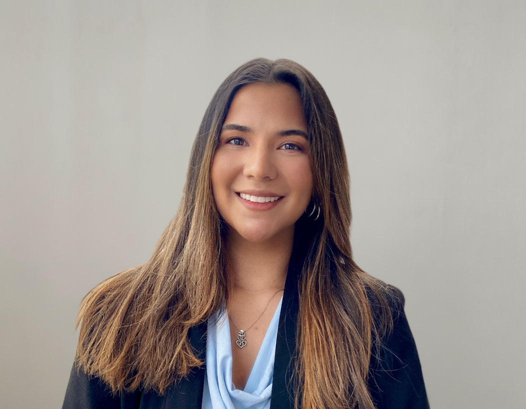A woman with long brown hair smiling, wearing a black blazer and a light blue blouse, standing against a plain light gray background.