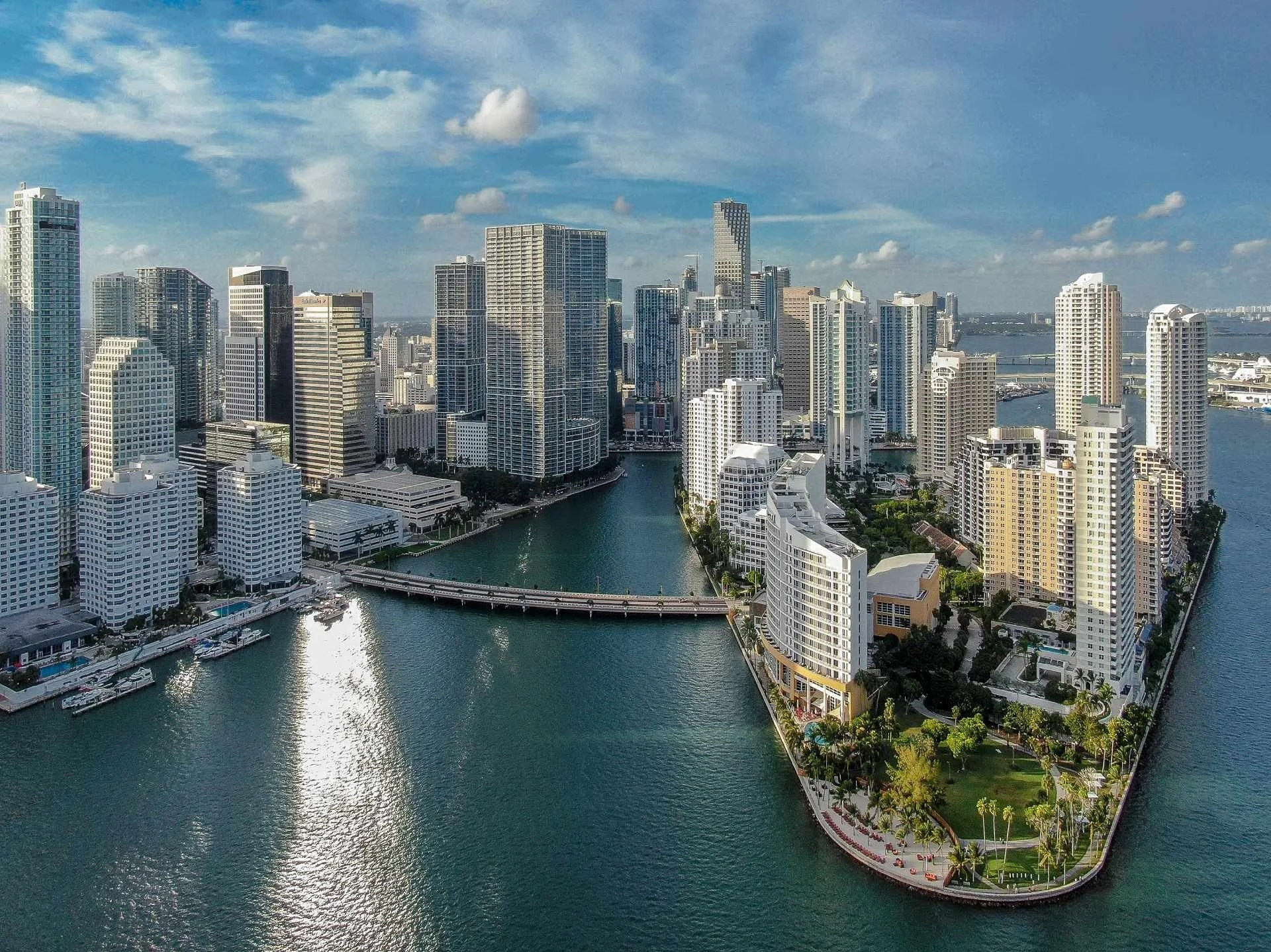 Aerial view of Miami FL skyline with high-rise buildings surrounding a waterway, featuring a curved beachside park with palm trees.