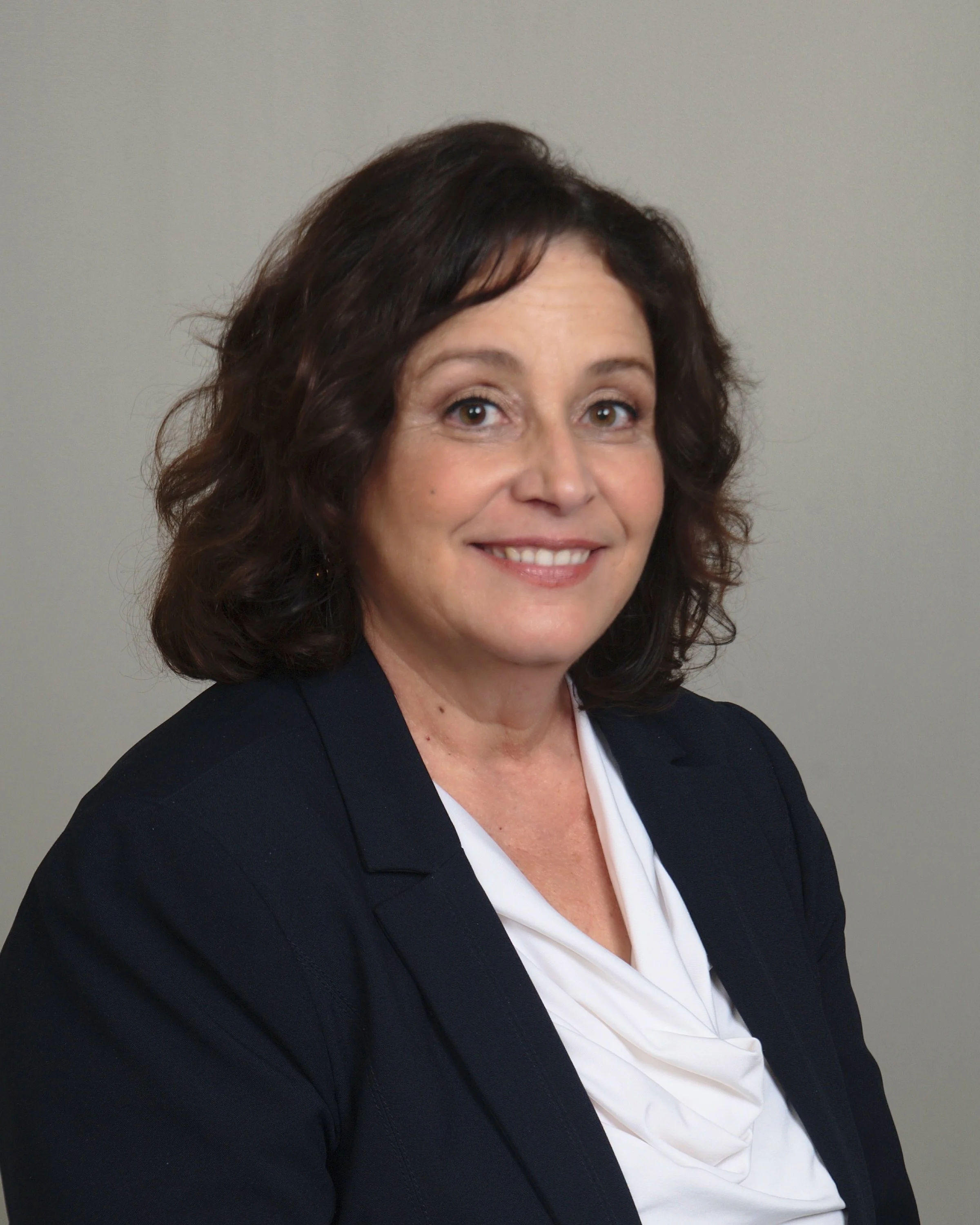 A smiling woman with dark brown shoulder-length hair, wearing a dark blazer and a white blouse, against a plain gray background.