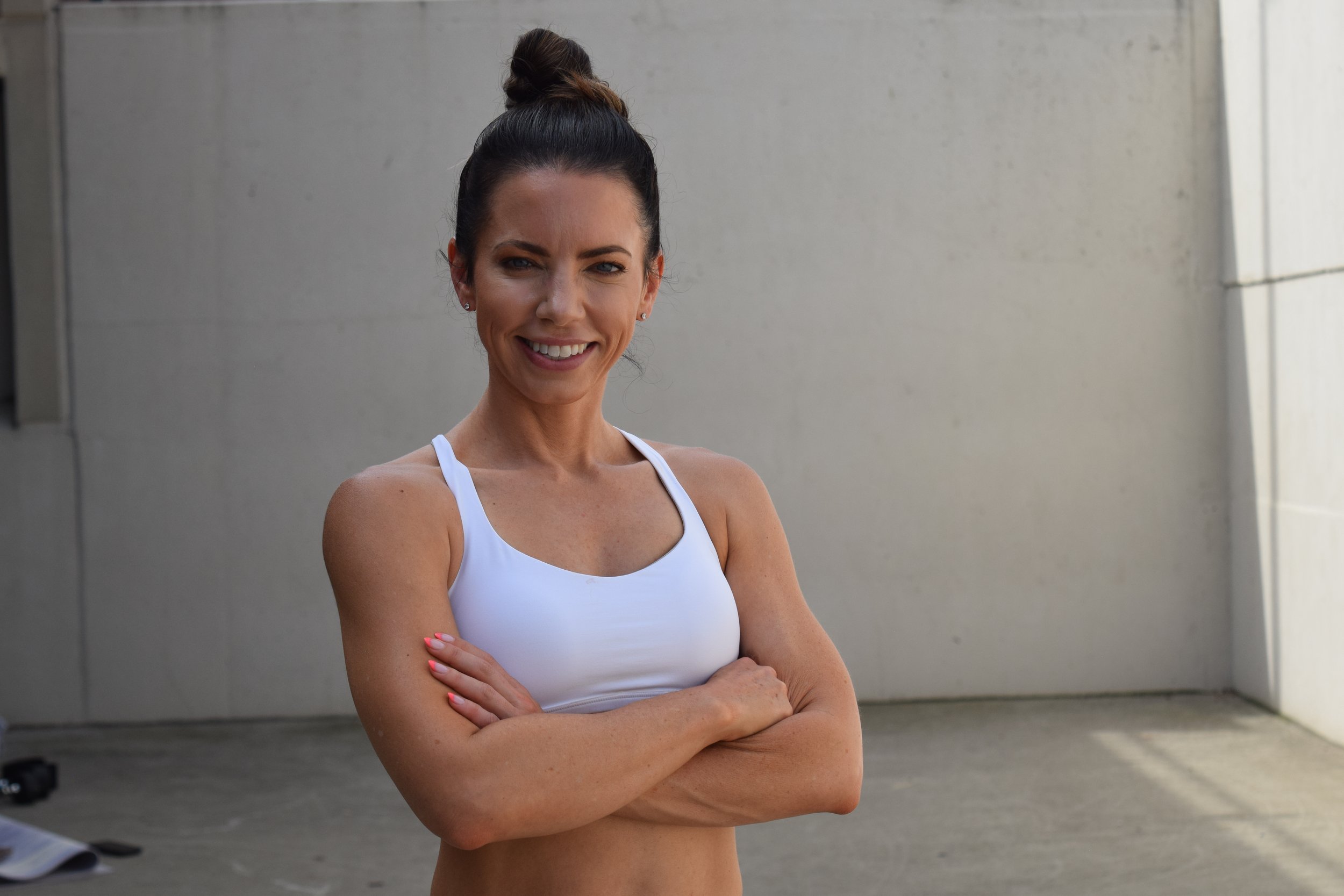 Smiling woman with cross arms in workout attire standing outdoors near a gray wall.