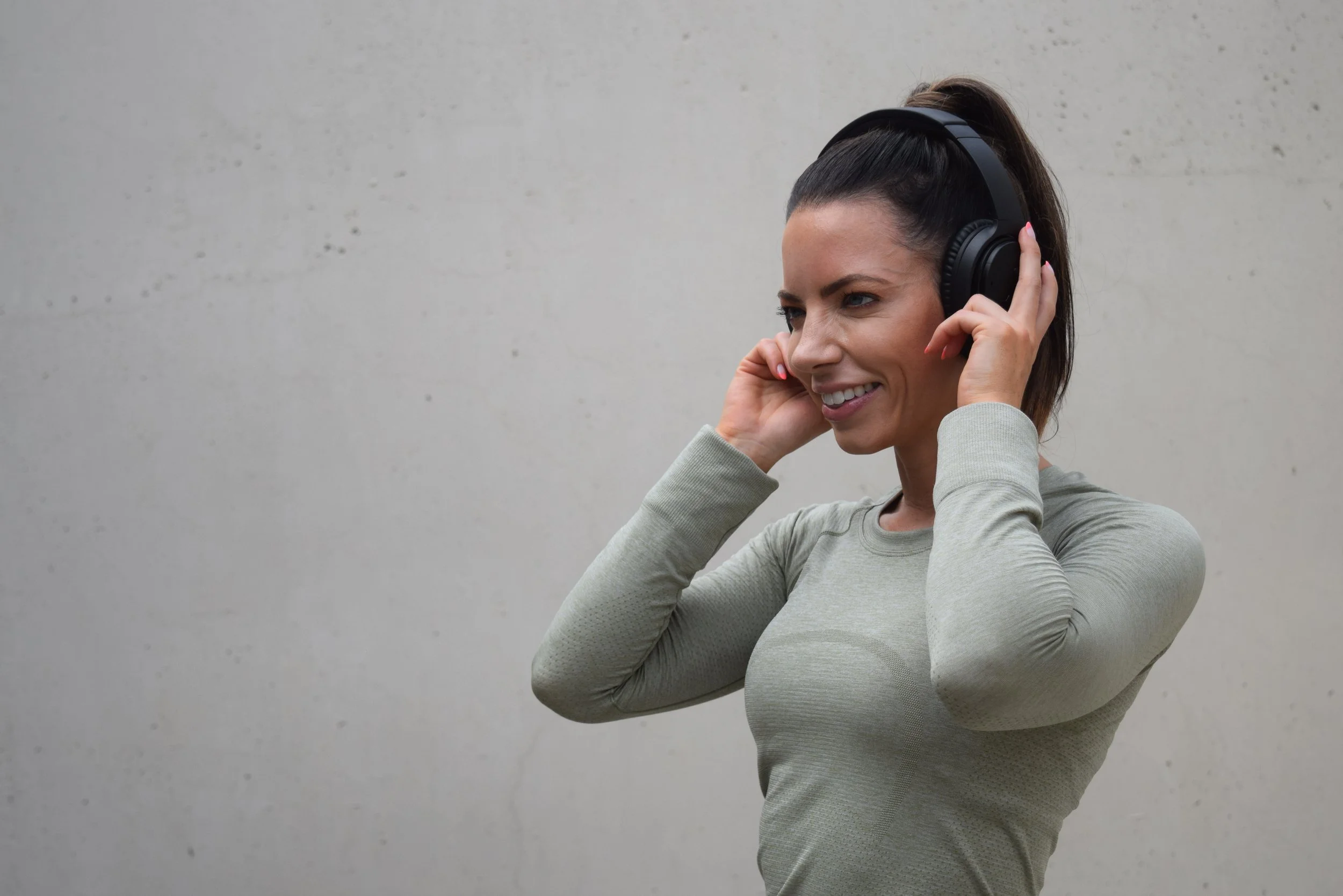 Woman, Ann Marie Wakula, in workout clothes listening to music with headphones, smiling against a plain wall.