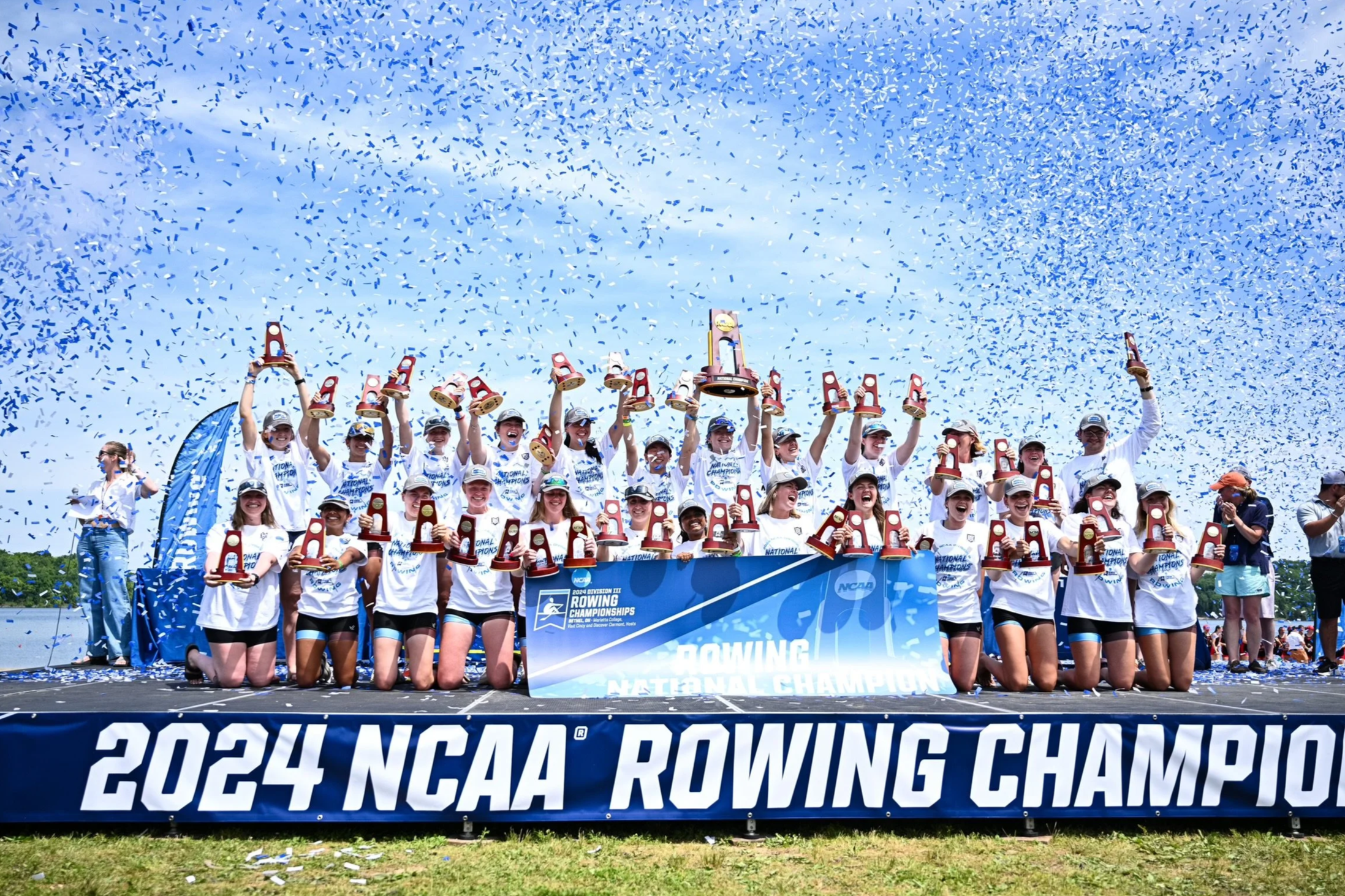 A team of female rowers celebrating their victory at the 2024 NCAA Rowing Championships, holding medals and trophies as blue confetti falls from the sky, with a blue backdrop and a large banner that reads '2024 NCAA Rowing Champions'.