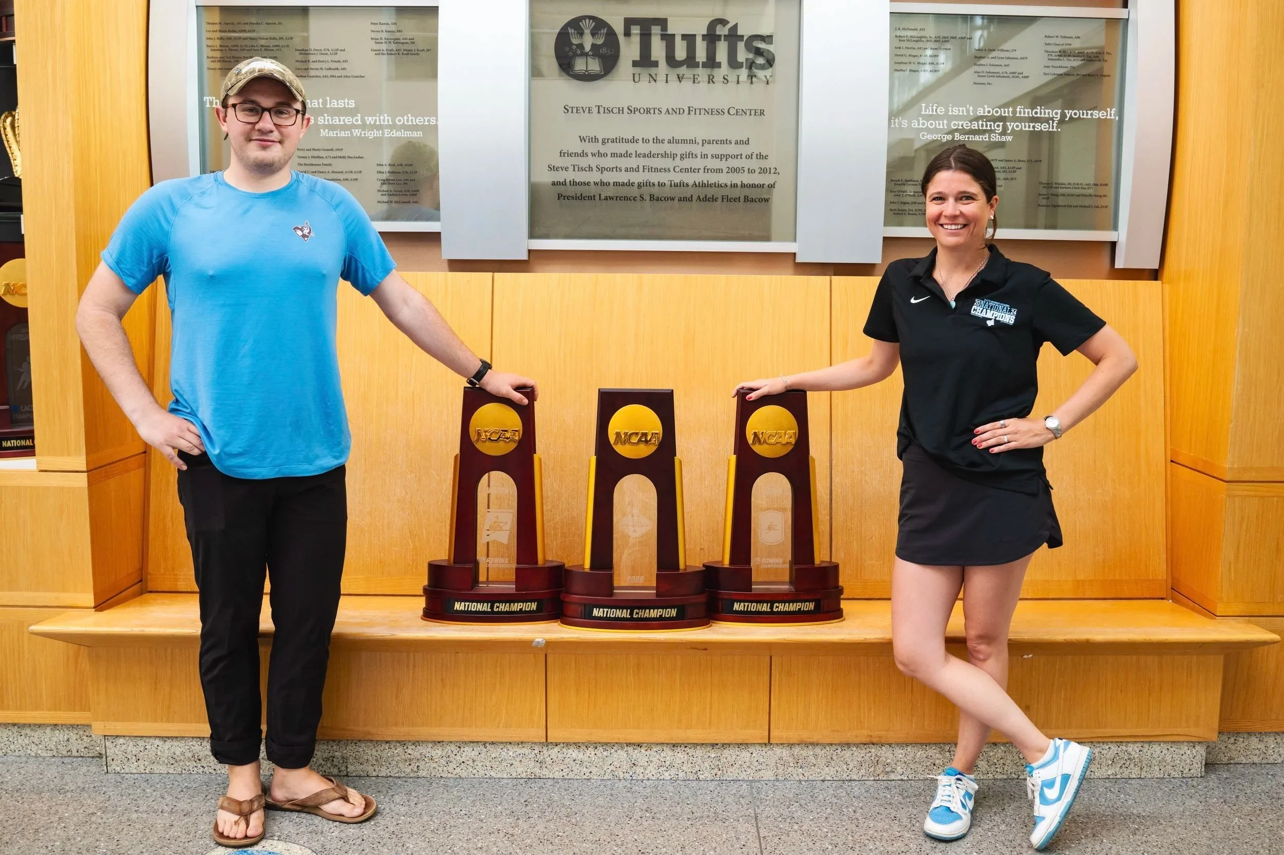 Two people, a man in a blue shirt and a woman in a black Nike shirt, standing in front of a display with NCAA trophies and a sign for Tufts University, smiling at the camera, with a wooden wall background.