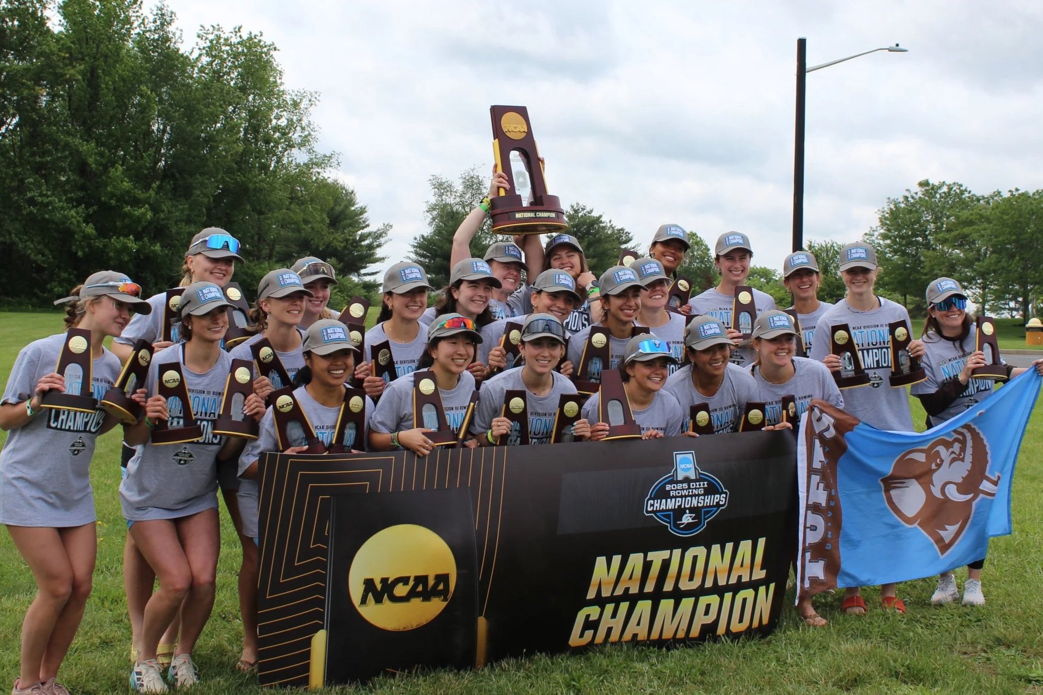 A women's rowing team celebrating their NCAA national championship victory, holding trophies, a banner, and a flag outdoors on a grassy field with trees and a cloudy sky in the background.