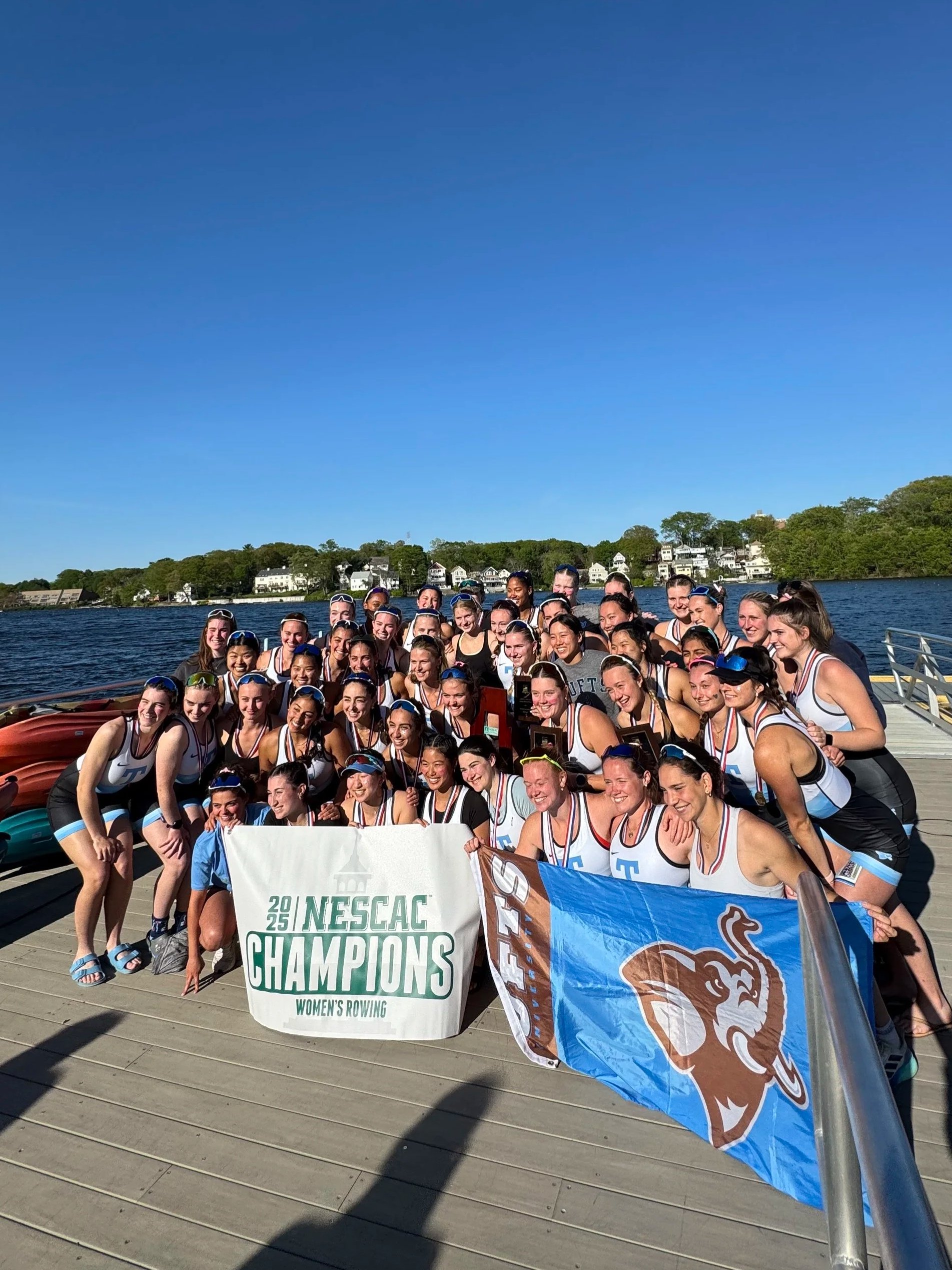 A large group of female rowers celebrating on a dock, holding banners that read 'NESCAC Champions Women's Rowing 2025,' with a body of water and houses in the background.