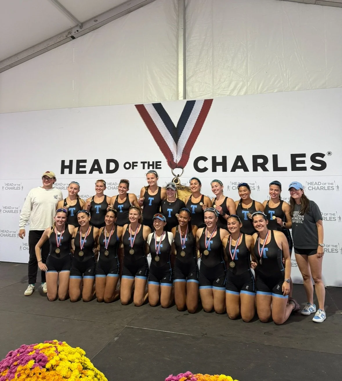 A girls' sports team with medals and coaches posing on stage at the Head of the Charles rowing race, with a large white backdrop featuring the event's logo and a medal hanging from a ribbon.