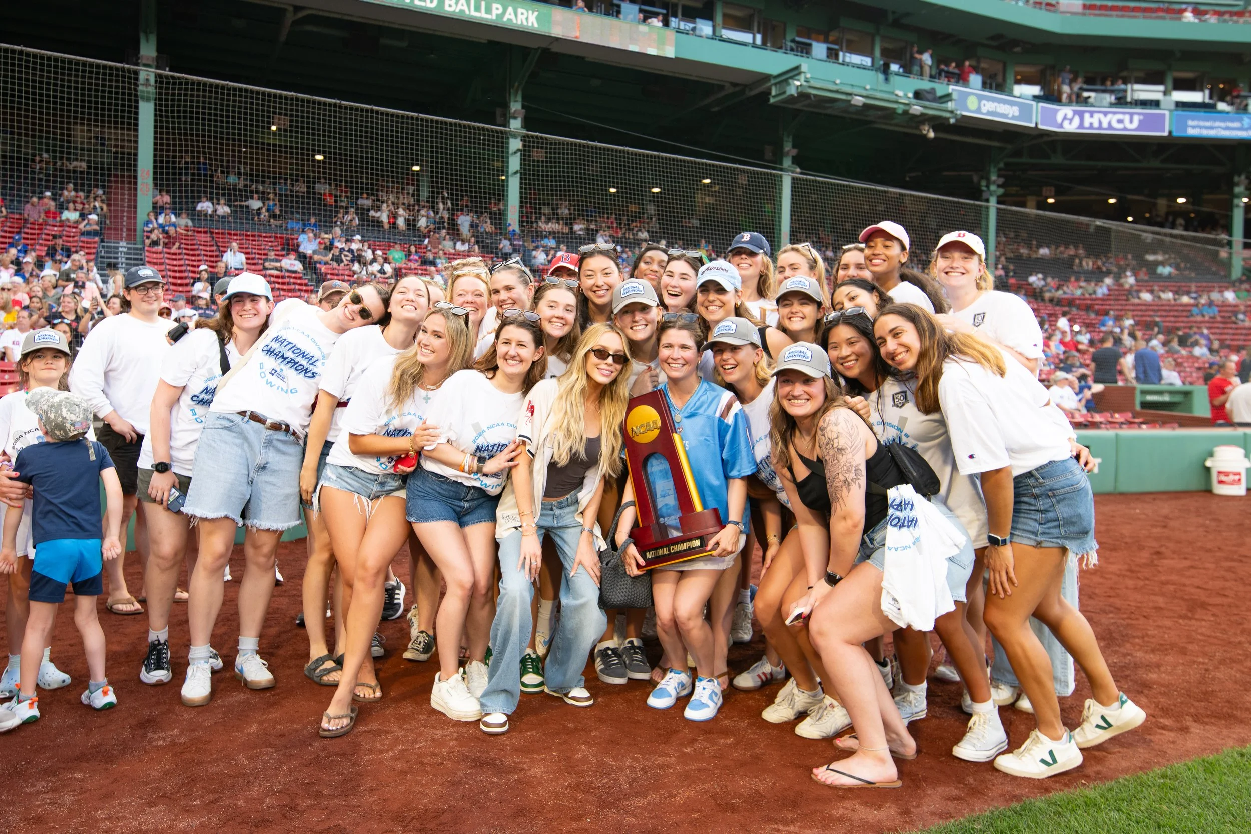 Group of young women celebrating their victory on a baseball field, holding a trophy, with many spectators in the stands.