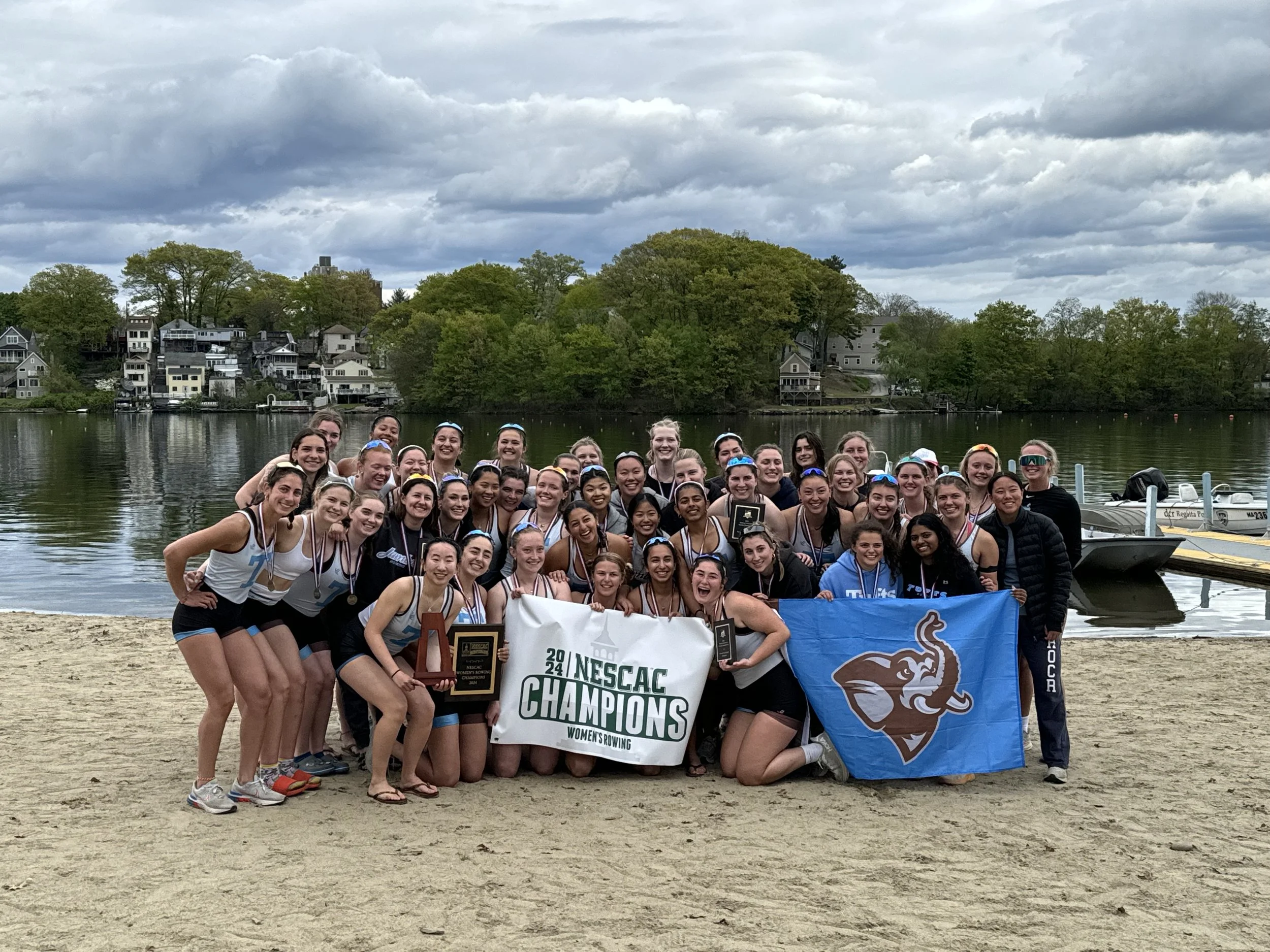 A large group of young women, who appear to be a rowing team, celebrate on a sandy beach by a lake. They are holding banners that read 'NESCAC Champions Women's Rowing' and display a blue flag with an elephant logo. Many are wearing medals and some are holding trophies, smiling and posing for the photo. In the background, there are boats docked near the water and a scenic view of houses and trees under a cloudy sky.
