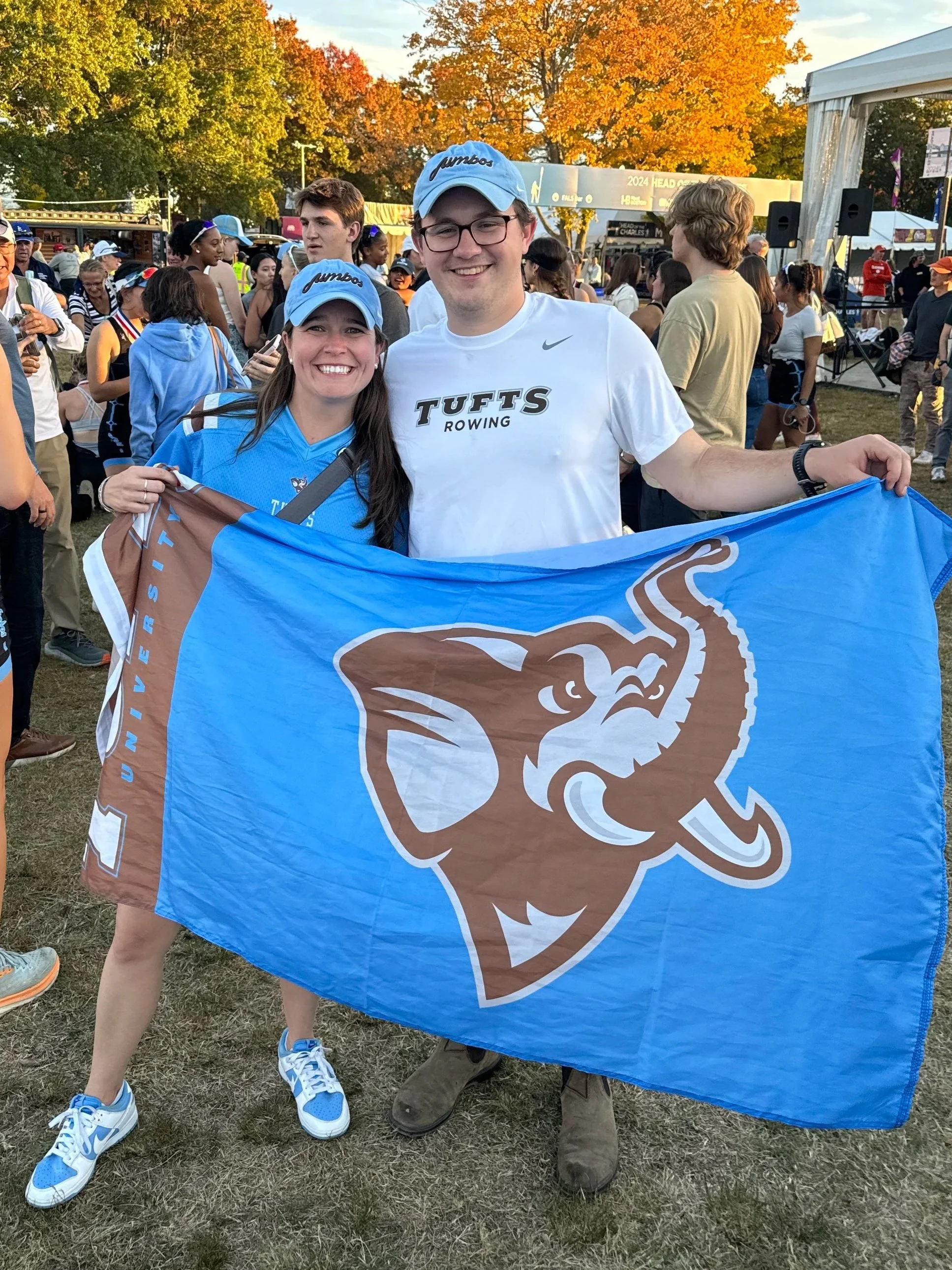 A smiling man and woman holding a Tufts University flag. They are at an outdoor event with many people, trees with autumn leaves, and a stage in the background. The woman is wearing a blue Tufts cap and a blue shirt, and the man is wearing a white Tufts rowing t-shirt and a blue Tufts cap.