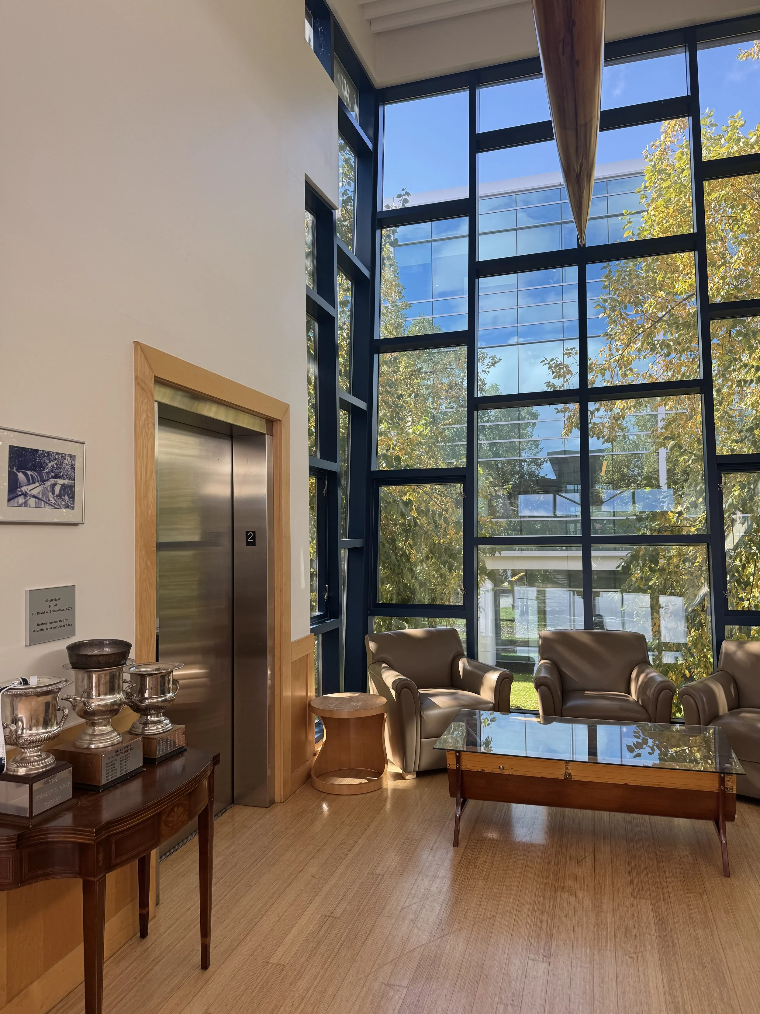 Modern lobby with large glass window wall, leather armchairs, a glass coffee table, an elevator, and trophies on a side table.