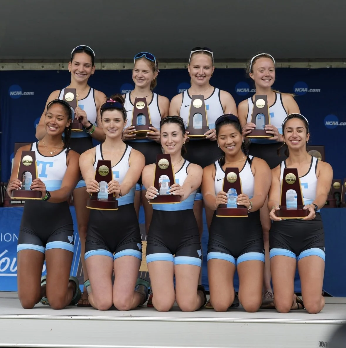 Nine young female athletes in matching black and blue uniforms smiling and holding NCAA trophies, kneeling in front and standing behind a blue podium at a sports award ceremony.