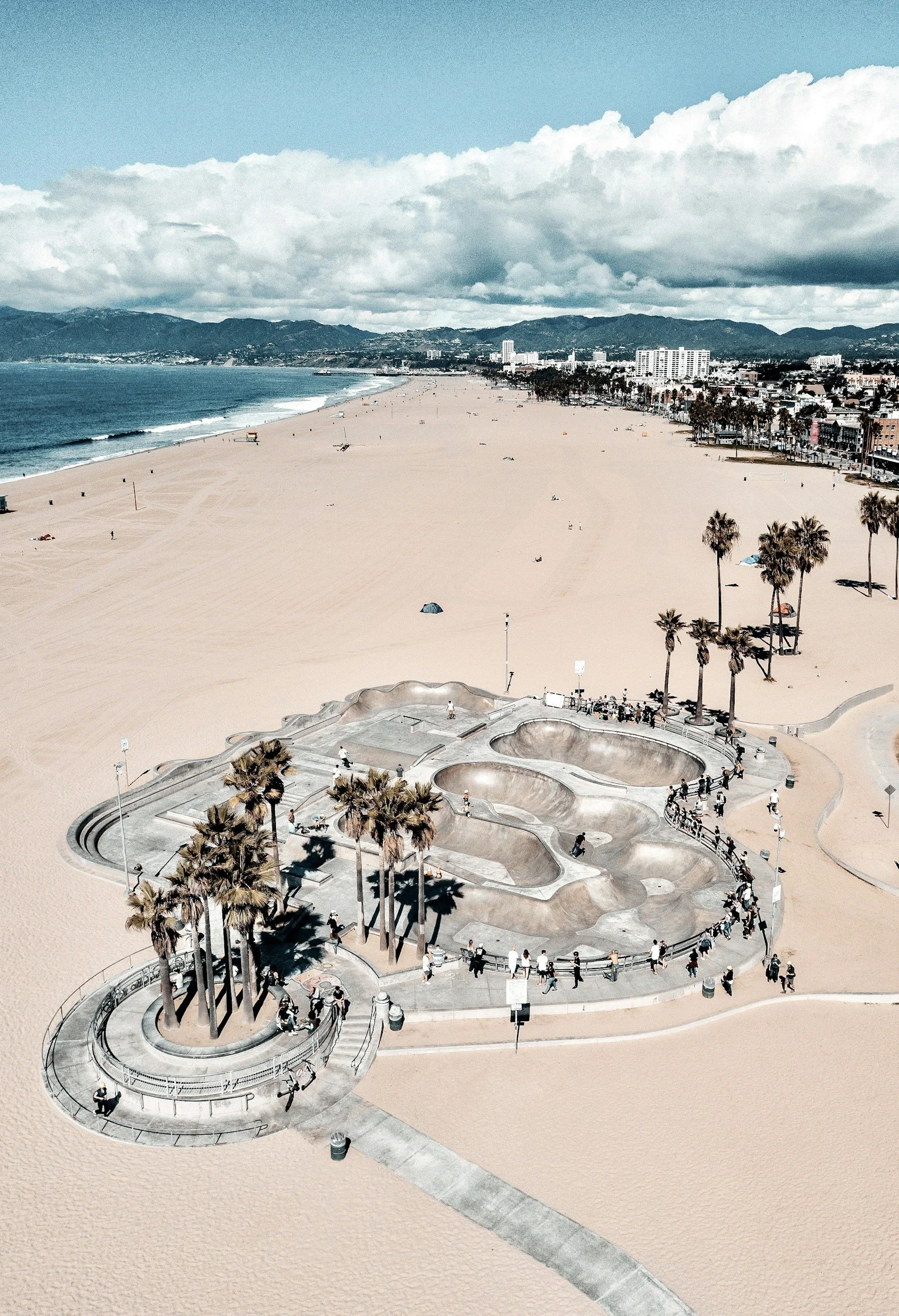 An aerial view of a sandy beach with a skate park and palm trees, with the ocean on the left and a cityscape with mountains in the background.