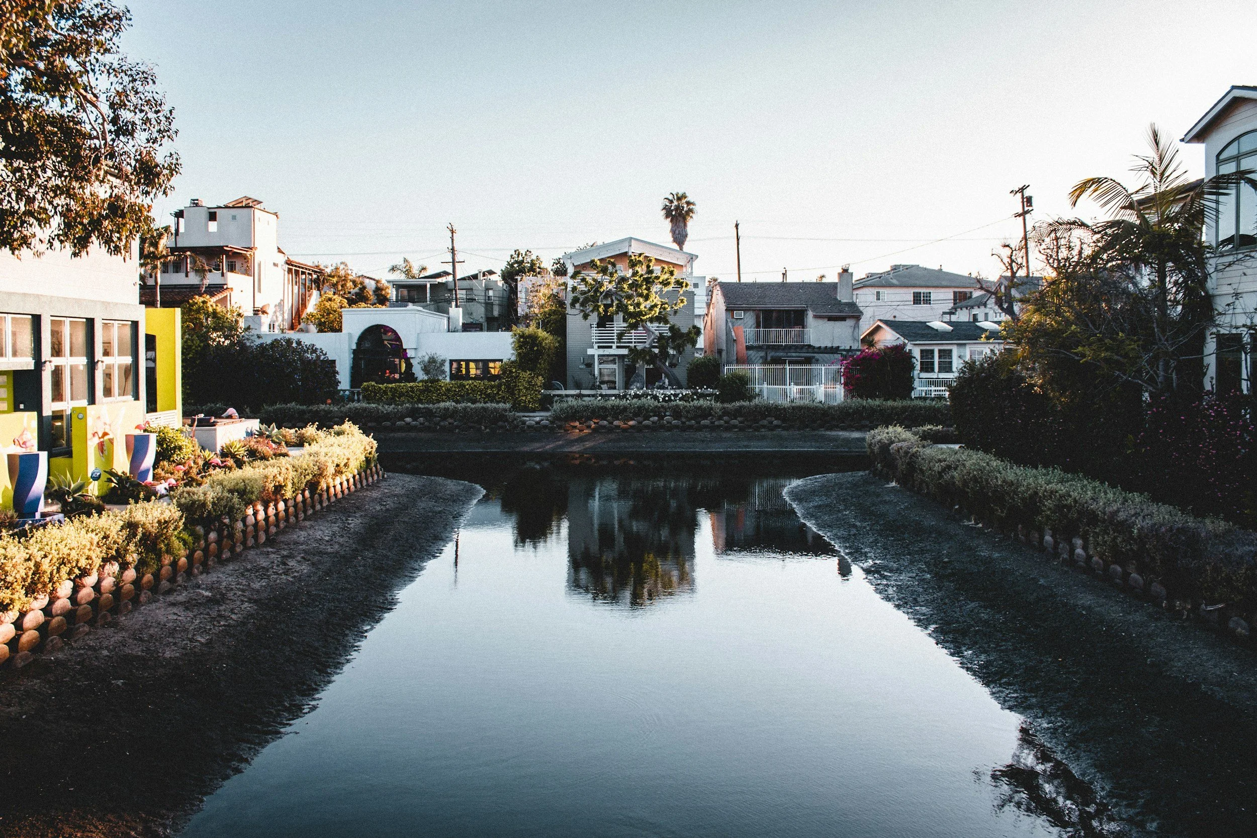 City houses with a small canal or pond in the foreground, trees, and clear sky.