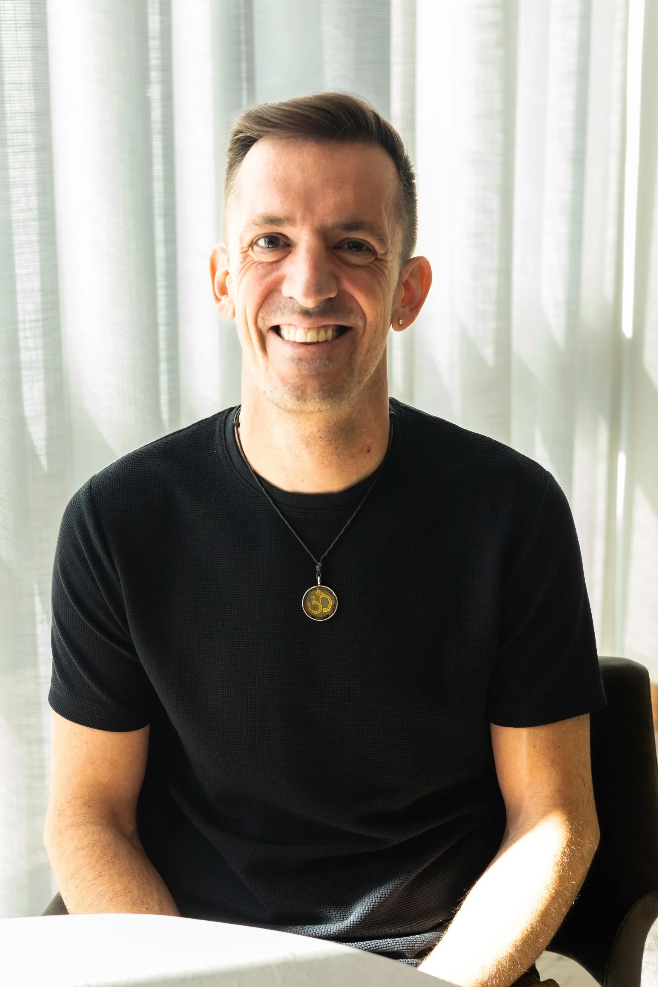 A smiling man with short brown hair wearing a black shirt and a necklace with a gold pendant, sitting indoors in front of light-colored curtains.