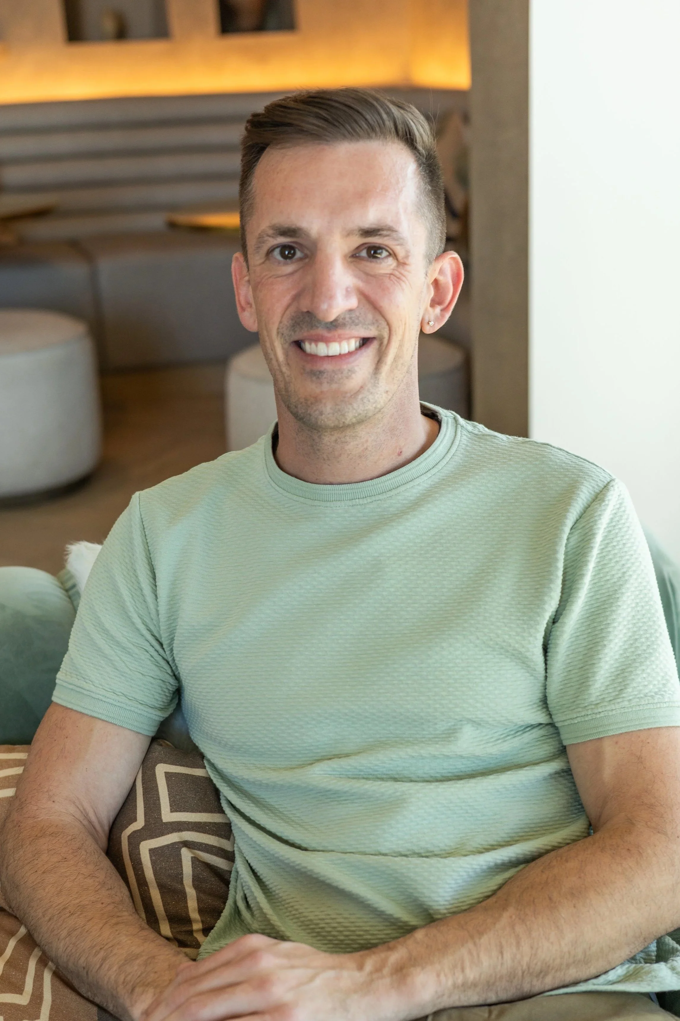 A smiling man with short brown hair is seated on a sofa in a room with warm lighting. He is wearing a light green t-shirt and has an earring in his left ear.