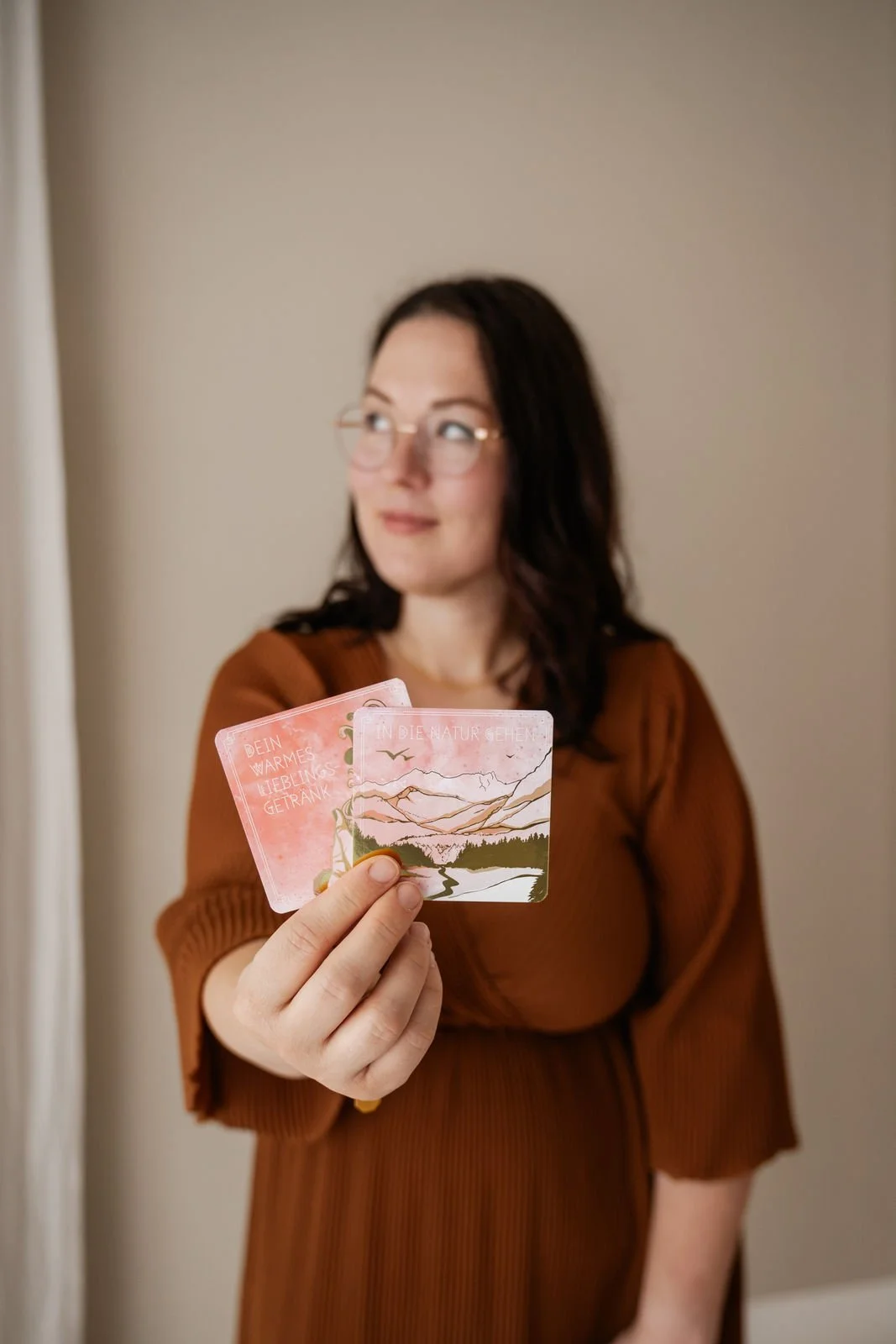 Eine Frau mit dunklem Haar und Brille hält eine Karte in die Kamera. Die Karte hat ein Naturbild mit Bergen, Fluss und Bäumen und Text in Deutsch.