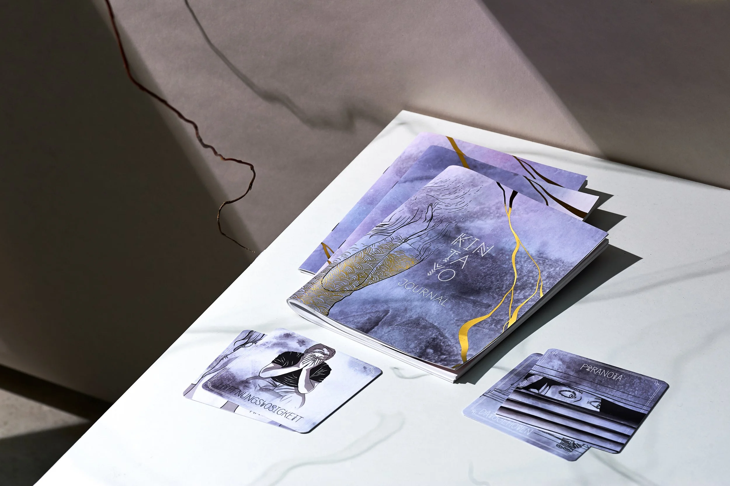 Ein aufgeschlagenes Notizbuch und zwei Karten auf einem weißen Tisch. Das Notizbuch hat einen blauen, lila und goldfarbenen Umschlag mit einer Berglandschaft. Eine Karte zeigt eine Illustration eines Menschen mit verschränkten Armen, die andere eine stilisierte Augenzeichnung mit deutschen Texten.