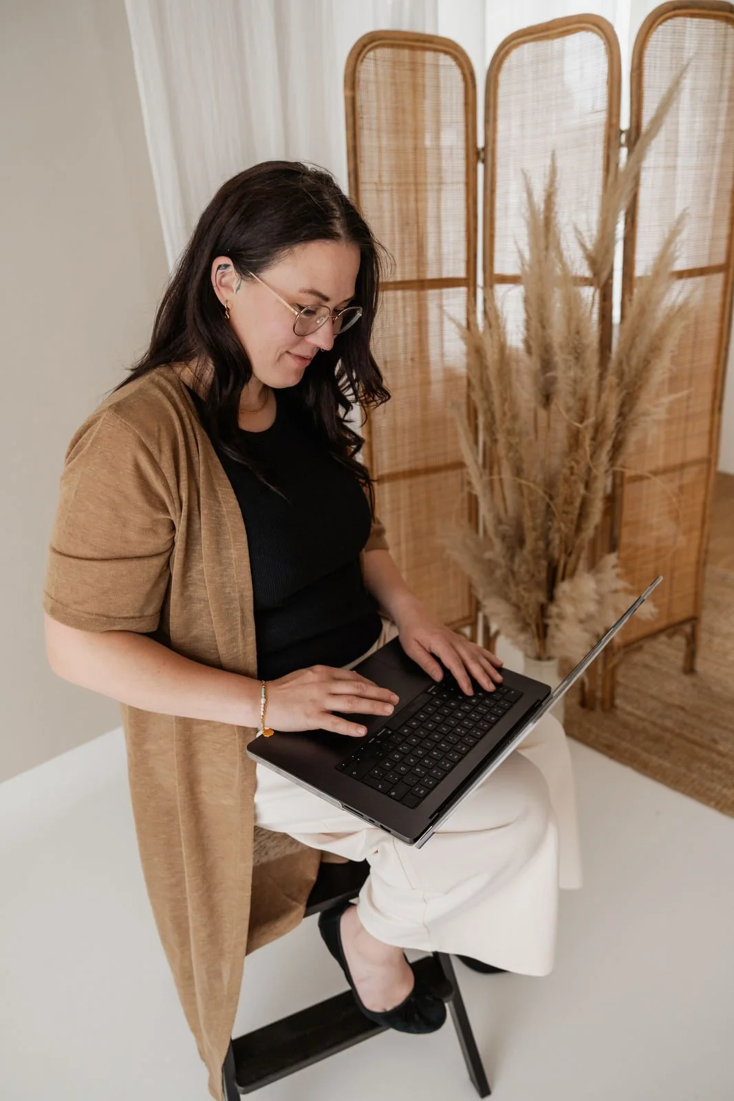 Frau arbeitet an einem Laptop in einem dekorativen Raum mit einer Trennwand aus Holz und Trockenblumen im Hintergrund.