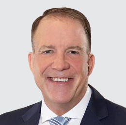 Portrait of a man in a suit with short brown hair, smiling against a light gray background.
