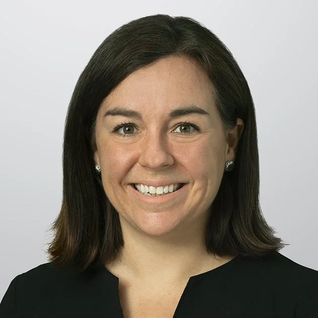 Headshot of a woman with shoulder-length dark hair, wearing earrings and a black top, smiling against a plain background.