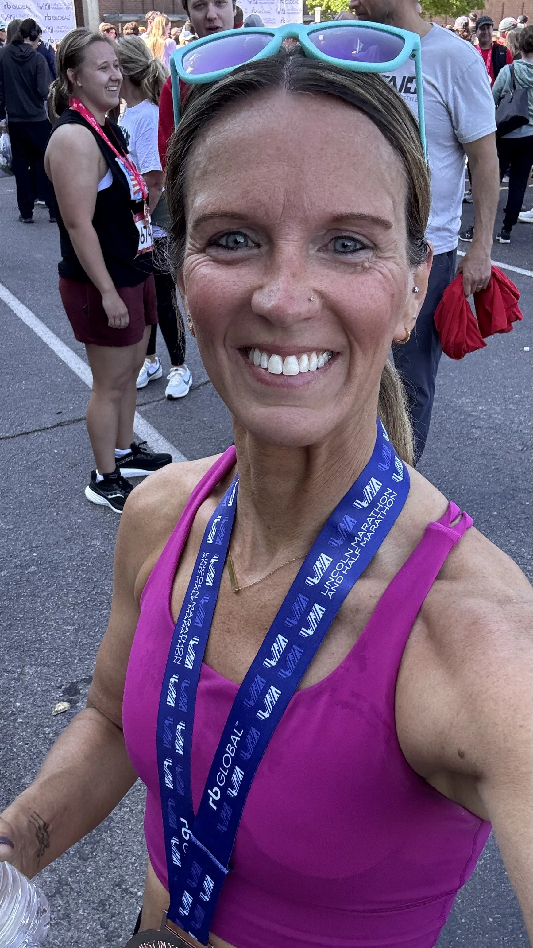 A smiling woman wearing a pink tank top and a medal around her neck, taking a selfie at a marathon event, with other runners and spectators in the background.