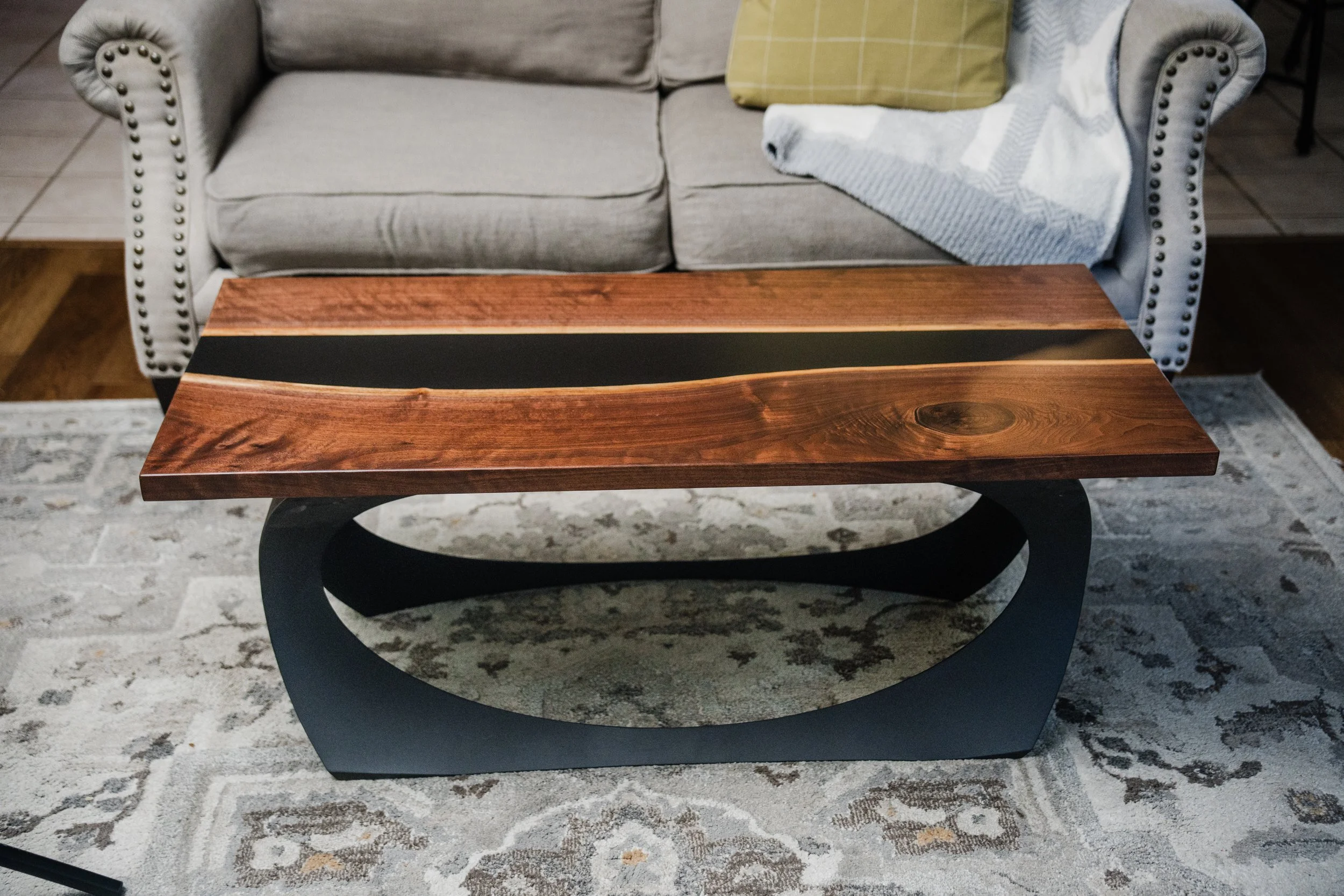 A wooden and black metal coffee table with a curved base, placed in front of a light-colored sofa with a yellow and a patterned throw blanket on it, on a patterned area rug.