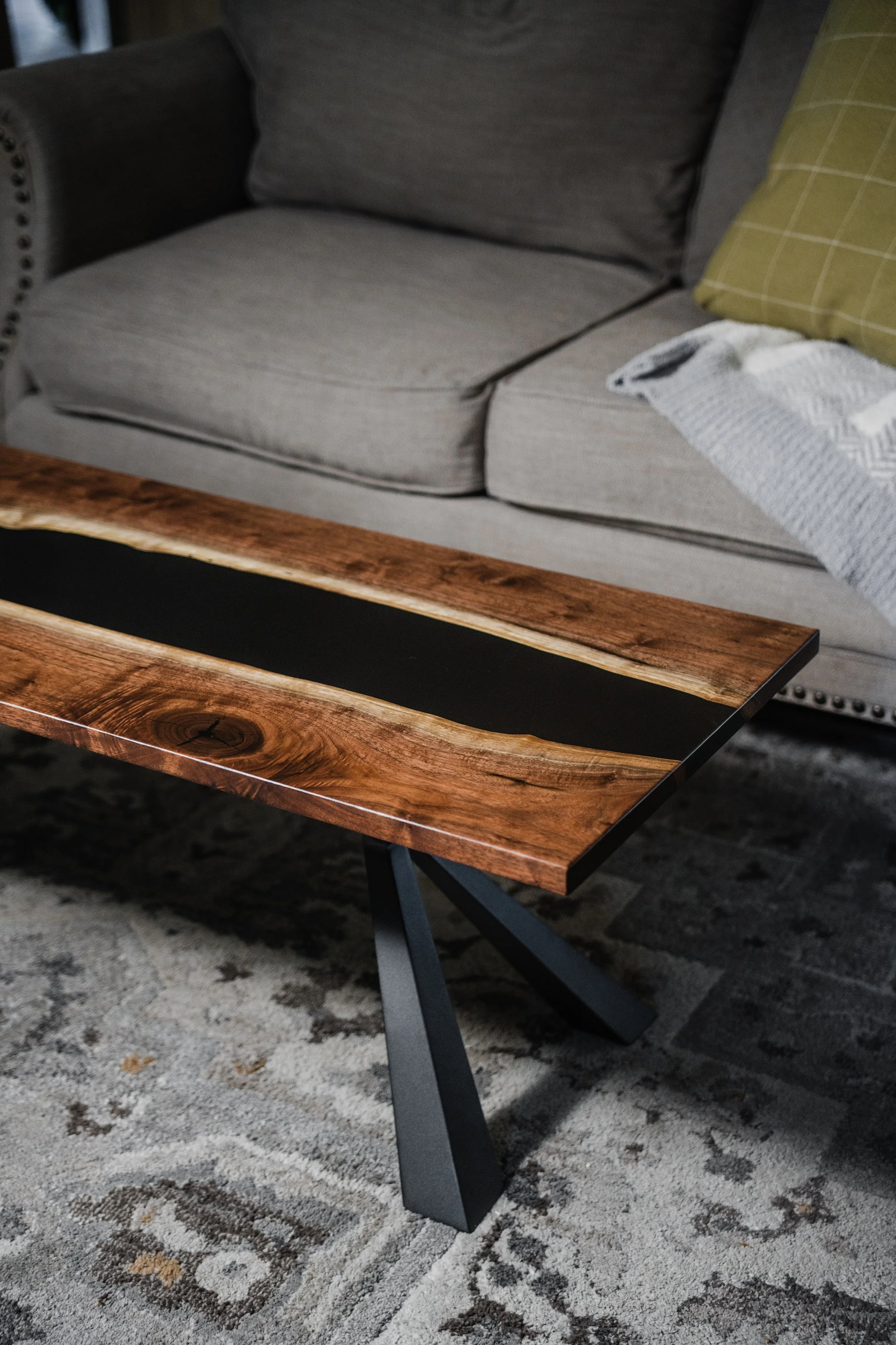 A coffee table with a wood top and black interior on a patterned area rug in front of a gray upholstered sofa with throw pillows and a blanket.
