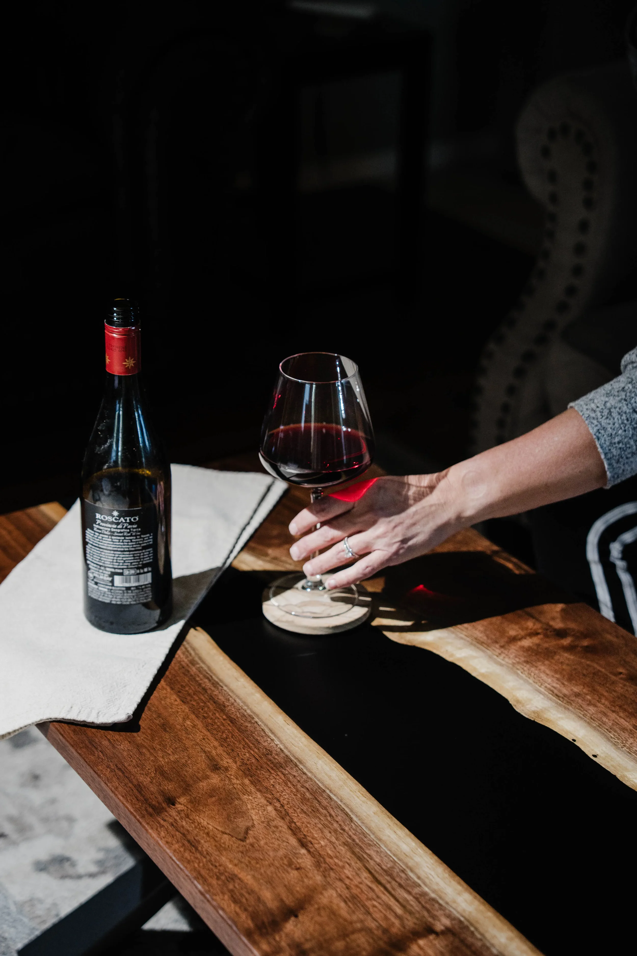 Person holding a glass of red wine next to a wine bottle on a wooden table.