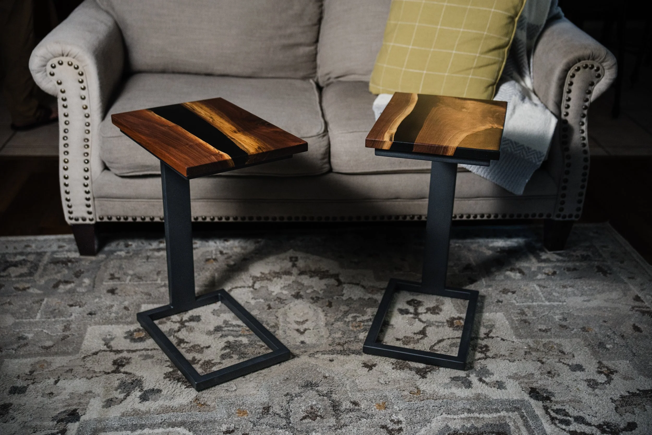 Two wooden side tables with black metal bases placed in front of a beige upholstered sofa with studded detailing, a yellow pillow, and a blanket on the armrest, on a patterned area rug.