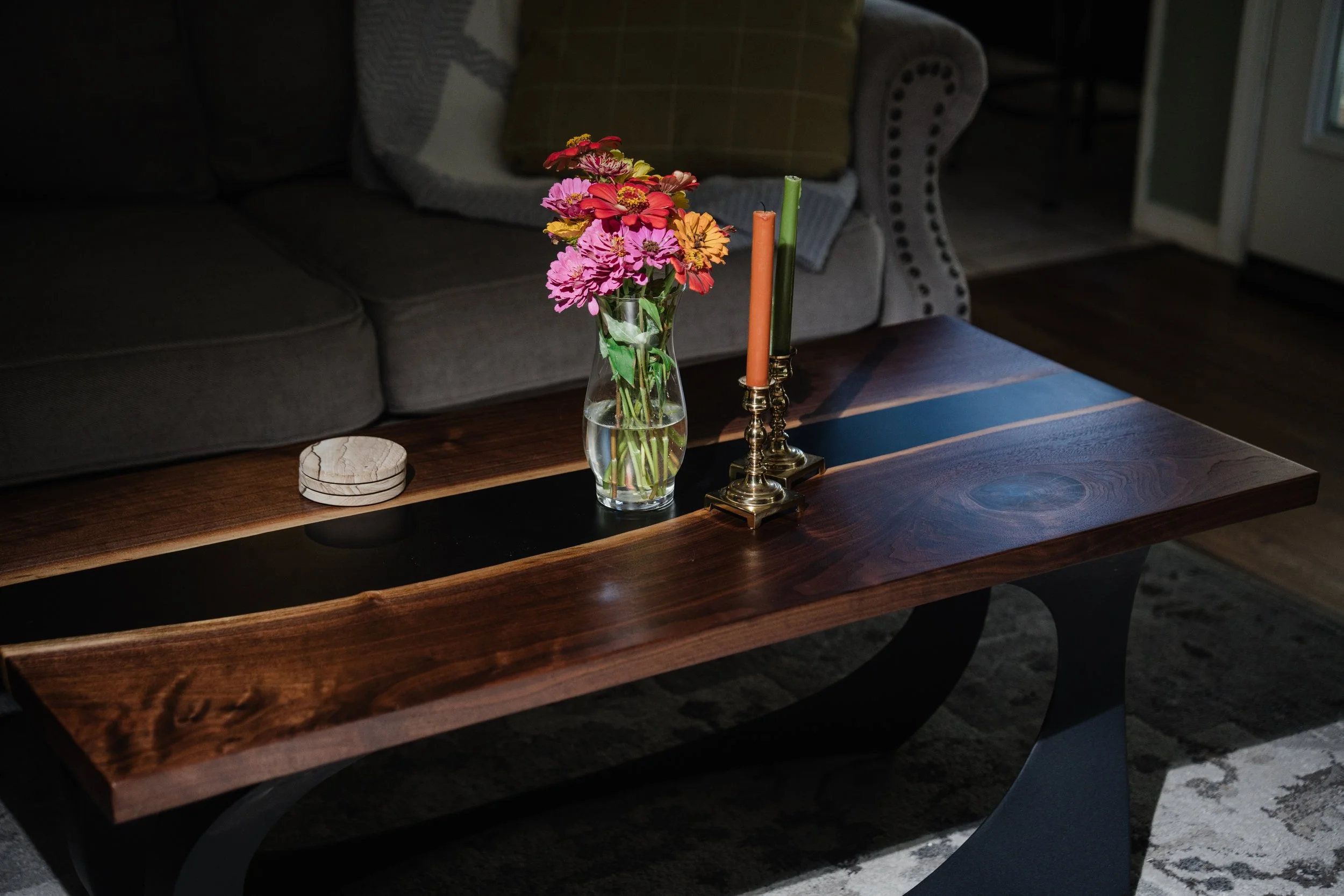 A wooden coffee table with a glass vase of pink, orange, and yellow flowers, two colorful candles in brass holders, a small round object, and a dark couch with pillows in the background.