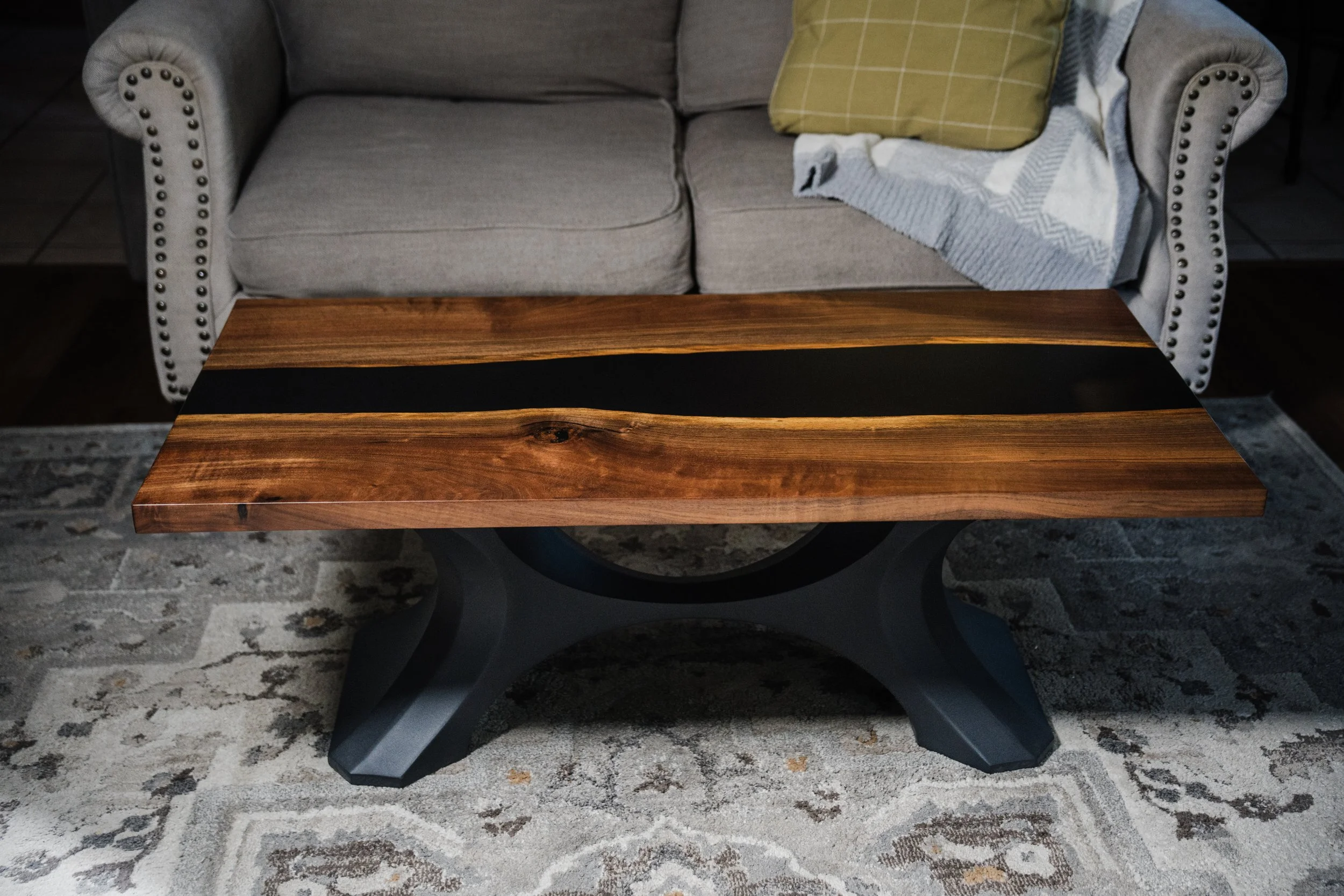 A wooden coffee table with a black stripe down the center, placed in front of a beige sofa with pillows and a blanket in a living room.