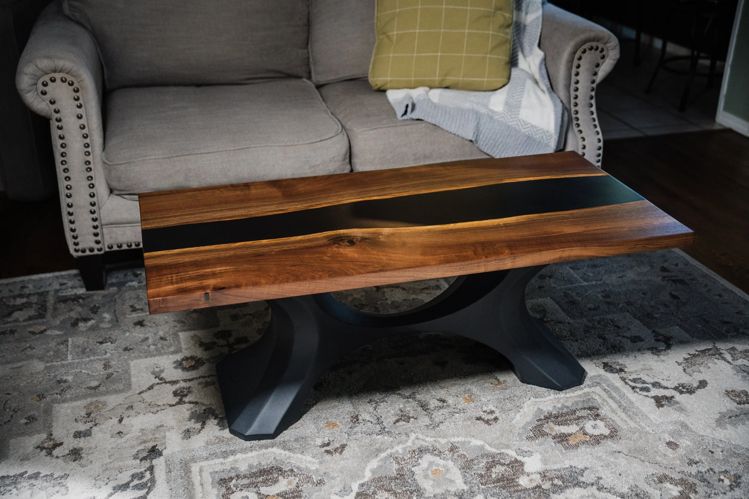 A wooden coffee table with black inlay in a living room setting, placed on a patterned rug in front of a gray upholstered sofa with studded armrests and a green pillow.